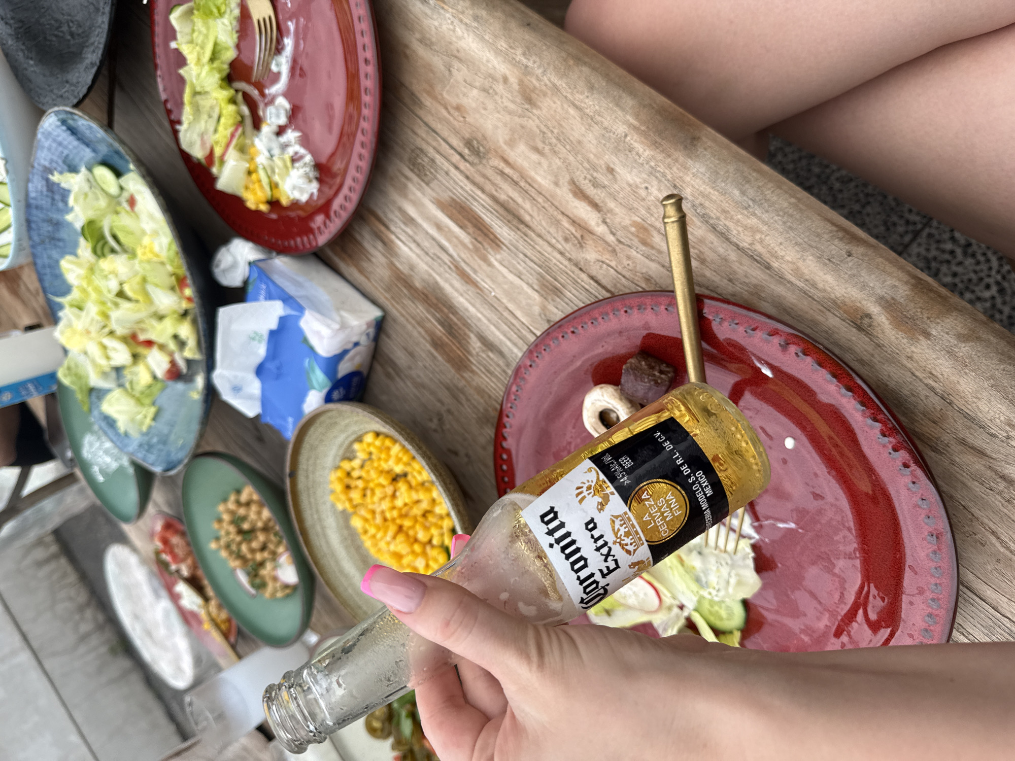 The image presents a top-down view of a casual dining scene, likely outdoors. A person's hands and lower arms are visible, one hand holding an empty "Coronita Extra" beer bottle. Several plates are arranged on what appears to be a wooden table. The plates contain various foods, including salads and corn. A box of tissues sits nearby. The presence of legs in the upper right suggests other people are present. The specific location in Jinhua, China, cannot be determined from the given visual information.