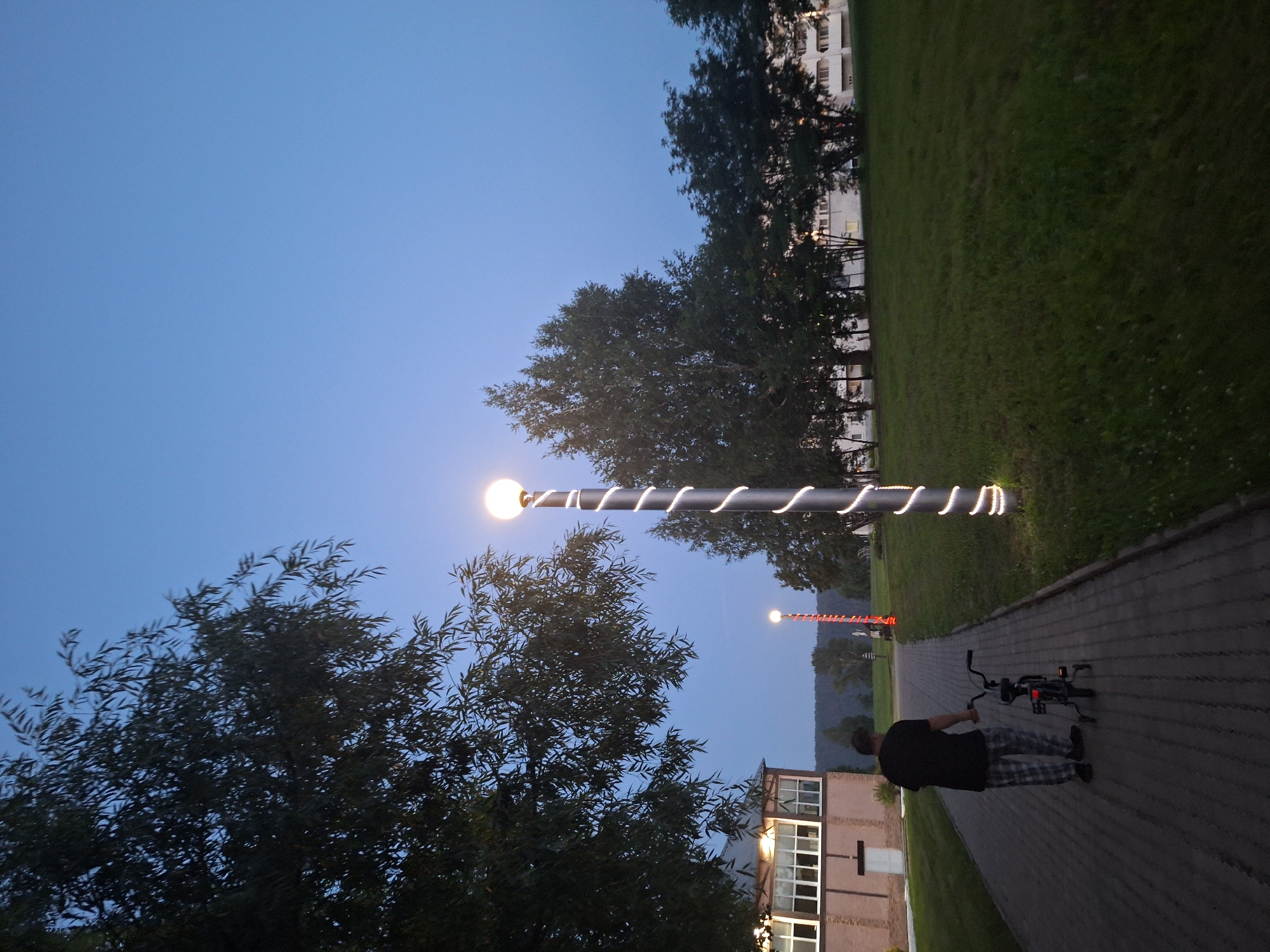 The image depicts an outdoor scene at dusk or early evening in Атамановка, Russia. A person wearing a dark t-shirt and checkered pants is walking on a paved pathway, pushing a small bicycle. The pathway is bordered by a grassy area on the right and a building with large windows on the left. Several trees are visible in the background, with a building complex further in the distance. Two lampposts are present; one in the foreground is tall and cylindrical with white spiral lights, and another is shorter and appears to be red with a spherical light at the top. The sky is a clear, deep blue. The overall environment suggests a residential or recreational area.