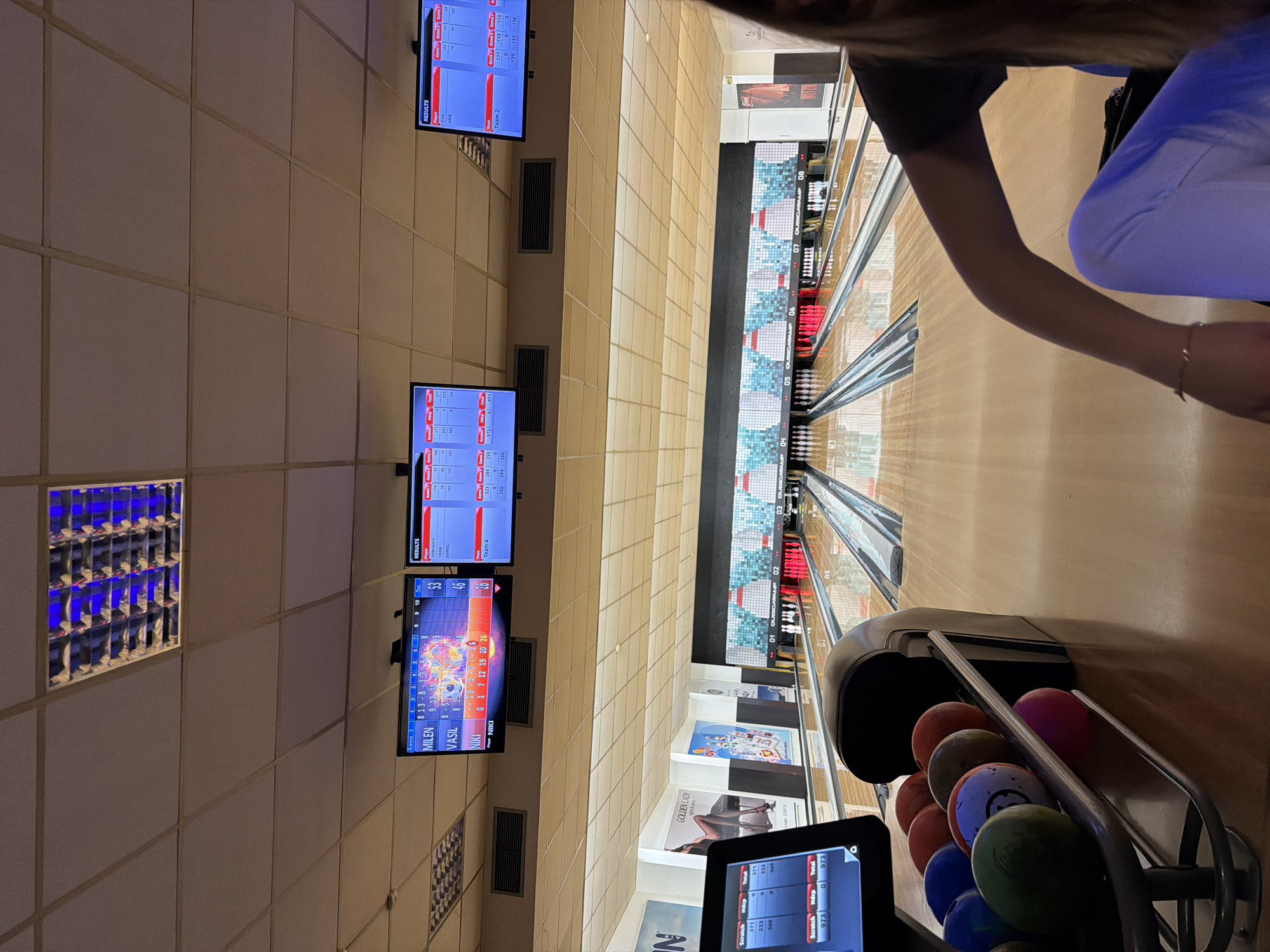 The image depicts the interior of a bowling alley located in Sofia, Bulgaria. The perspective is from the player area, looking down multiple bowling lanes toward the pins.
In the foreground, on the left, a metal bowling ball return rack holds several multi-colored bowling balls, including red, green, blue, orange, and pink. To the immediate left of the ball rack, a black-framed touchscreen display shows "Scratch," "Hdcp," and "Total" scores for Team 1 and Team 4. On the right, partially visible in the frame, is an individual seated, wearing a dark short-sleeved top and light blue/white pants. Their right arm, adorned with a delicate bracelet, rests on their lap, and long hair is visible on the far right.
Above the bowling lanes, three large display screens are mounted. The left screen shows player scores for "MILEN" (53 points), "VASIL" (46 points), and "NIKI" (26 points), with Niki currently designated as the player. The middle screen displays "RESULTS" for "ADRIYANA" (371 total), "YOAN" (233 total), and "DANIEL" (369 total), with a "Team 4" entry. The right screen also shows "RESULTS" for "EVA" (156 total) and "KRIS" (102 total), with a "Team 2" entry.
Eight bowling lanes are visible, sequentially numbered 01 through 08, with "QUBICAAMF" branding visible above some numbers. The lanes are constructed of polished wood. Bowling pins, some standing and some toppled, are visible at the end of the lanes; some are red and white. The walls behind the pins feature various advertisements, including a "GOLDEN LADY" poster depicting a woman's legs and another poster displaying text including "2,99 лв," indicating Bulgarian currency. The ceiling consists of light-colored acoustic tiles with integrated rectangular fluorescent light fixtures, one of which in the upper left corner emits a distinct blue light. The overall environment is brightly lit by artificial light.