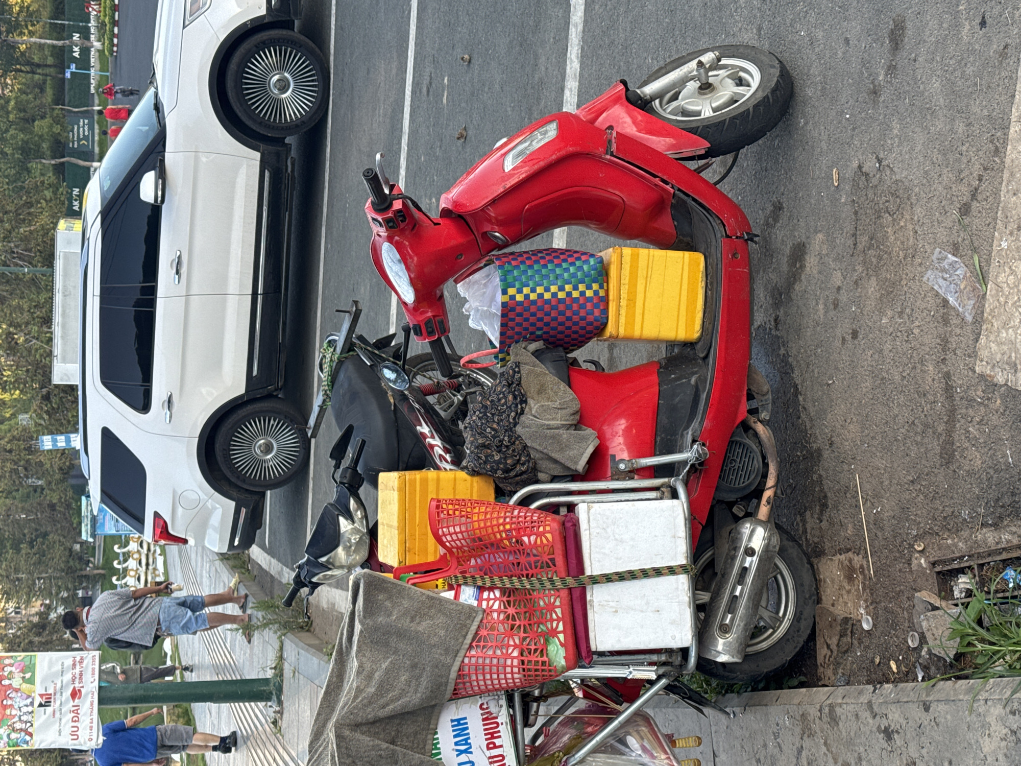 The image shows a red scooter parked on a street in Da Lat, Vietnam. The scooter is loaded with yellow containers and red baskets. A white SUV is parked next to the scooter. Two people are standing on the sidewalk near a green sign that reads "ƯU ĐÃI HỌC SINH SINH VIÊN 91 148 BA THÁNG HAI 1800 3355". The sidewalk is littered with trash.