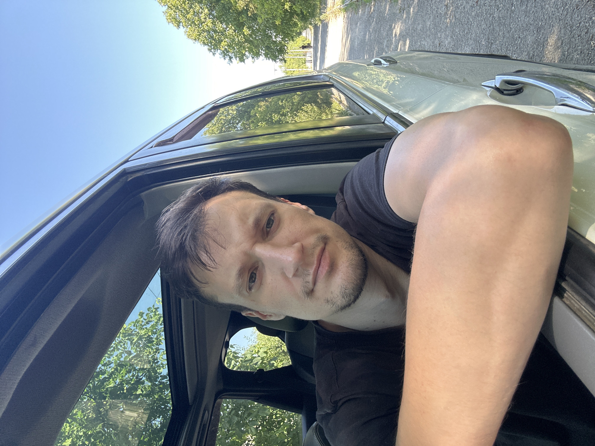 A man with short brown hair and a beard is leaning out of a car window, looking at the camera. He is wearing a black t-shirt. The car is light-colored and appears to be parked on a dirt road lined with trees. The sky is blue and clear. The image is a selfie taken from inside the car. There is a person walking in the distance on the road. The location is in the city of Makiv, Ukraine.
