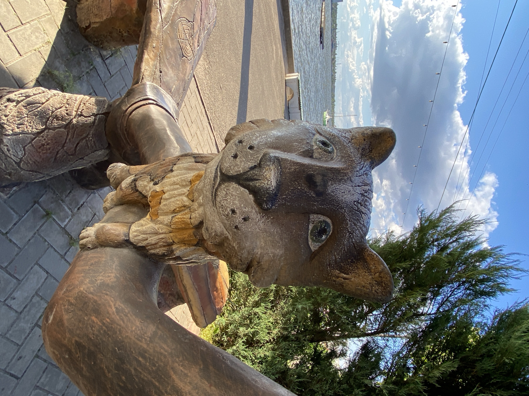 A carved wooden sculpture of a cat's head sits on a wooden bench outdoors. The cat sculpture has a brown and tan color palette, with visible wood grain and textured details. Its eyes are large and round, and its ears are pointed. The bench appears to be made of weathered wood, with carved patterns on its surface. The sculpture and bench are positioned on a paved area, likely a walkway, with a body of water and a distant cityscape visible in the background. Trees and vegetation are also present in the scene, with a bright blue sky and white clouds overhead.