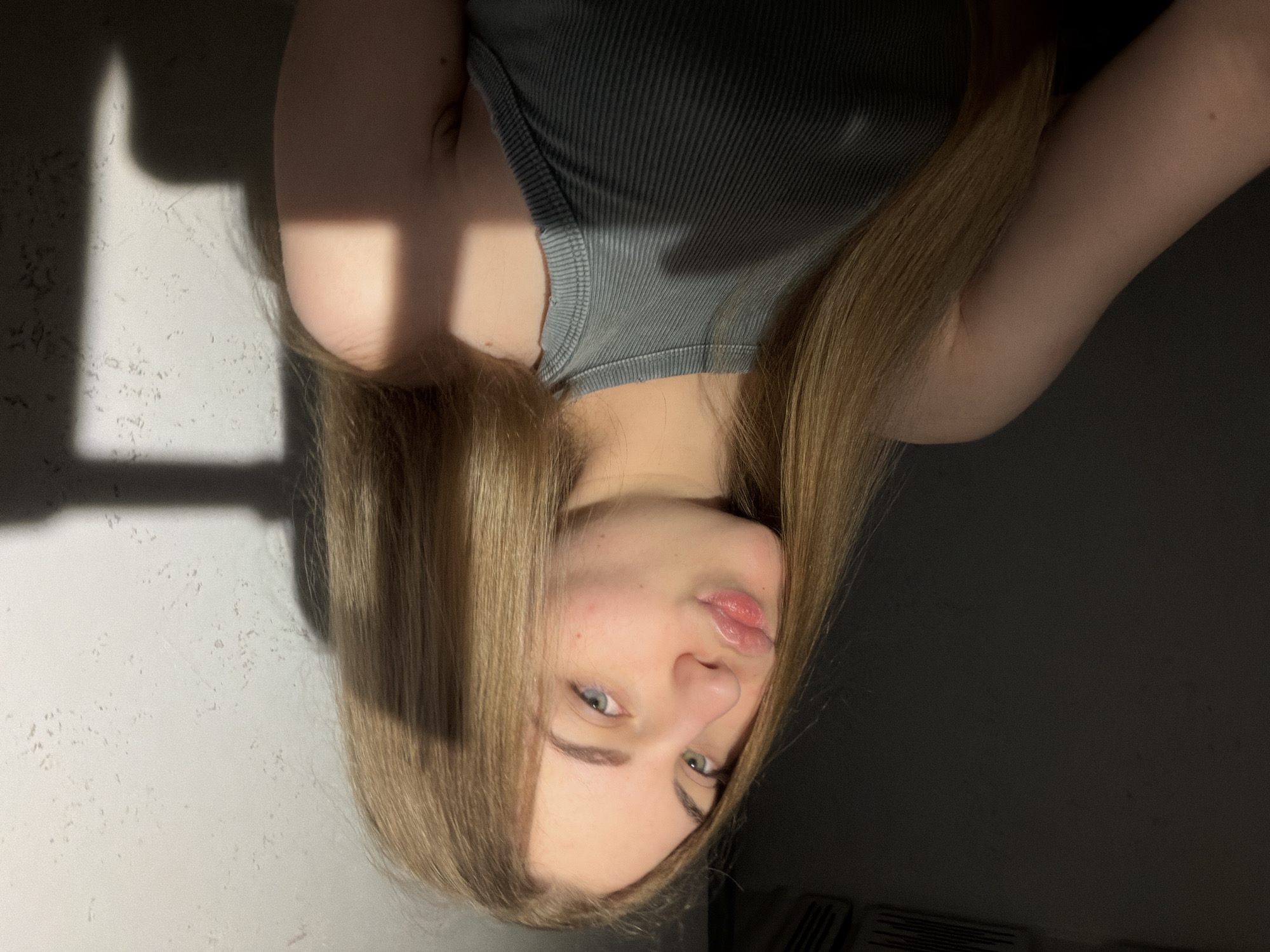 A young woman with long blonde hair and blue-green eyes is captured in a close-up shot, leaning into the frame. She is wearing a grey, ribbed tank top. Her lips are pursed in a playful gesture. The lighting suggests a warm, natural light source, casting a distinct shadow across the textured wall behind her, indicating an indoor setting, possibly a residential apartment. The play of light and shadow on the wall, and the woman's skin, suggests the sun is shining through a window or other aperture. The background is dark and indistinct on the left side of the frame, with the right side showing a textured light-colored wall. There is no specific identifiable information in the image that would pinpoint its location in Dnipro, Ukraine, or any other city.