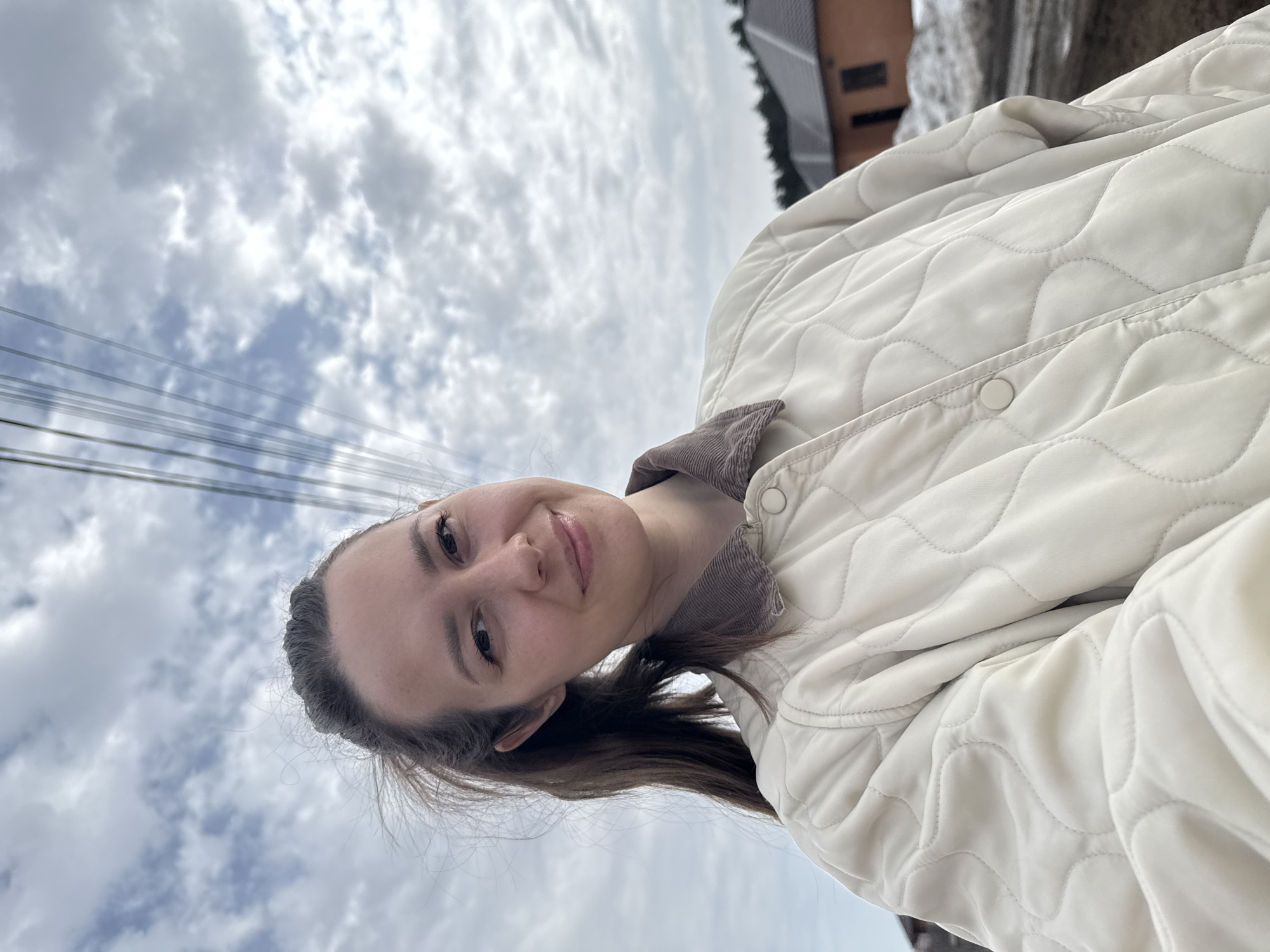 A woman is facing the camera, wearing a quilted beige jacket with a brown corduroy collar. The background consists of a cloudy sky, power lines, a building with a brown roof and white facade. The photo was likely taken outdoors. There is insufficient information to determine if the location is in the city of Chaikovsky, Russia.