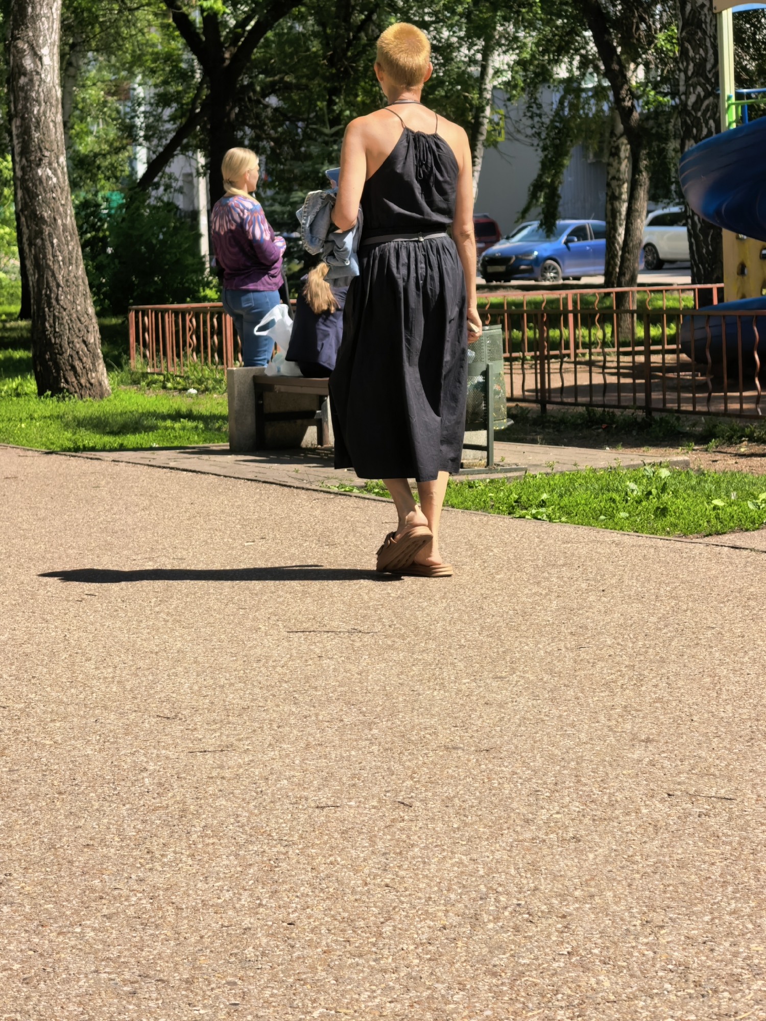 A woman with short, blonde hair, wearing a black halter dress and tan platform sandals, walks away from the camera on a paved path. She is holding a light-colored bundle in her left arm. In the background, a woman with blonde hair pulled back in a ponytail, wearing a purple and blue patterned sweater and blue jeans, sits on a bench. Next to her, a person in a dark blue shirt is visible. Behind the bench, there is a metal fence with vertical bars. Further in the background, several cars are parked, including a blue sedan and a white sedan. Trees and green grass are visible throughout the scene, suggesting an outdoor park or public space. The scene is well-lit, indicating daytime.