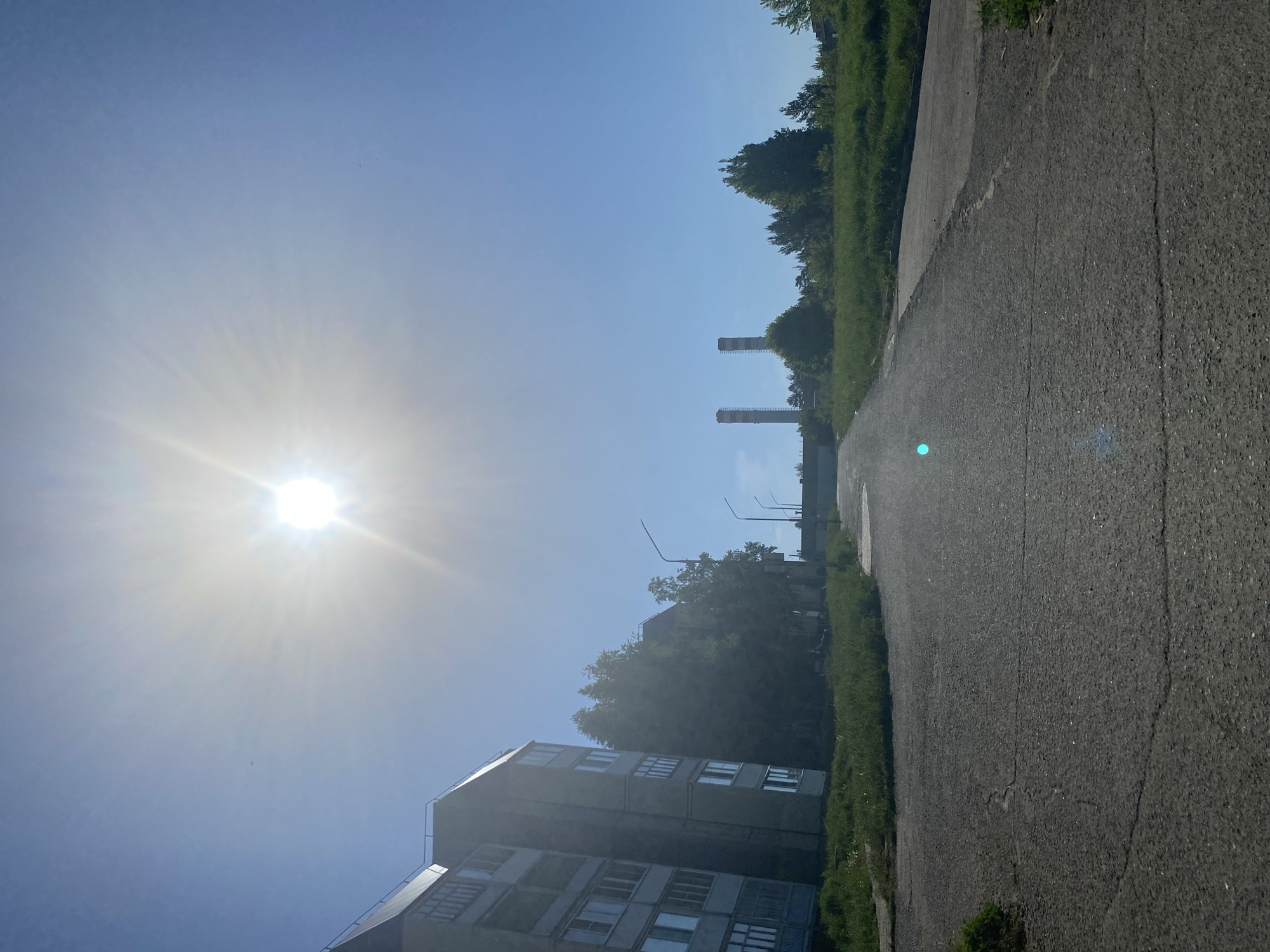The image shows a street in Марковский, Russia, on a bright, sunny day. The sky is clear and blue, with the sun positioned high in the frame. A tall, grey apartment building stands on the left side of the street. Along the right side is a paved road with grassy areas bordering it. Two industrial chimneys are visible in the distance, rising above the treeline. The lighting conditions suggest it is daytime, casting shadows and creating a glare in certain areas.
