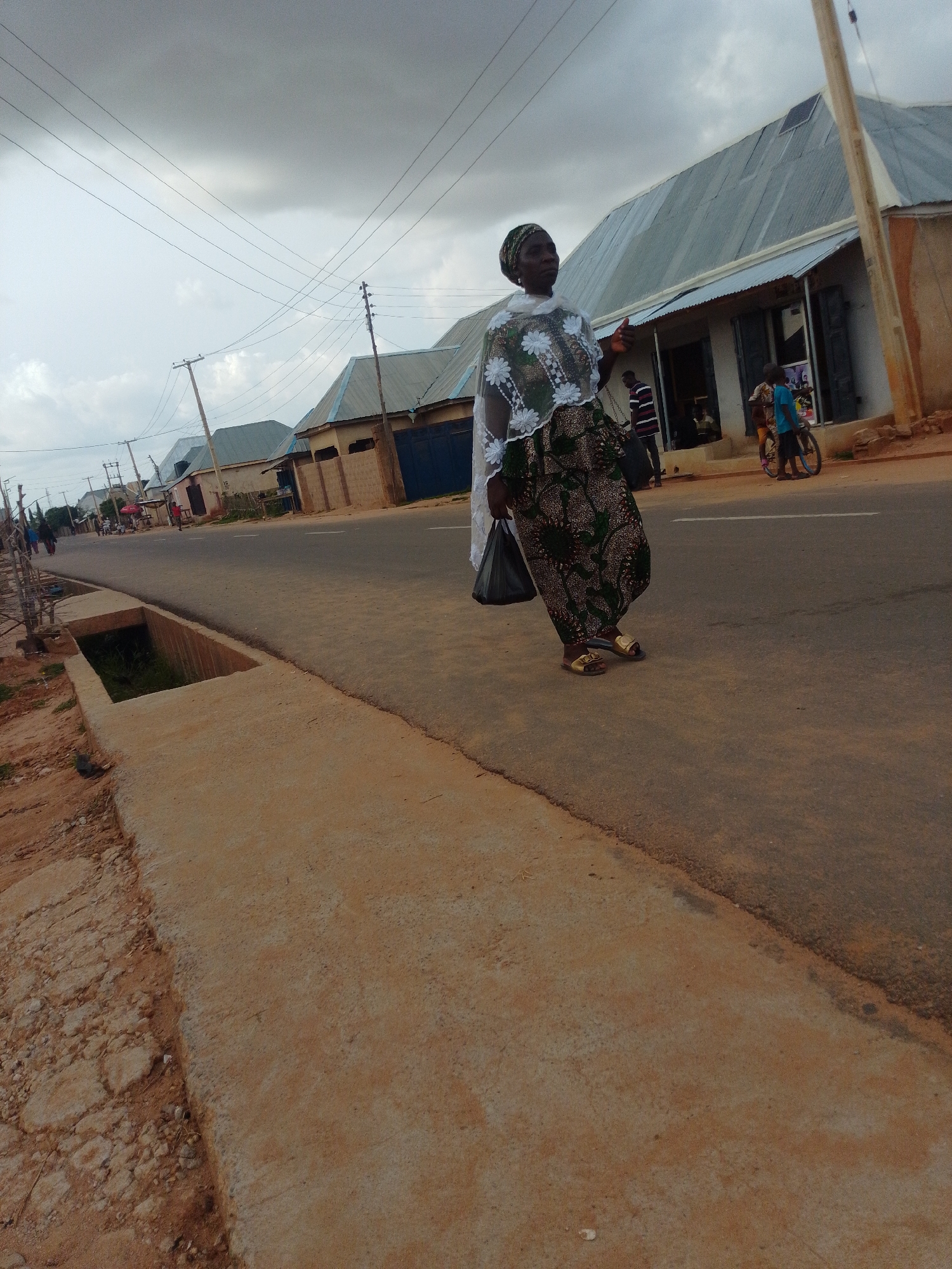A woman walks along a paved road in Tempure, Nigeria. She wears a green and brown patterned dress with a white, sheer overlay adorned with white floral embroidery, and a matching headscarf. She carries a black plastic bag in her left hand and wears gold sandals. The woman is walking on the right side of the road, facing away from the camera, towards a row of buildings. The buildings have corrugated metal roofs and appear to be shops or residences. A child on a bicycle is visible in front of one of the buildings, and another person is seen behind the woman, also walking away from the camera. The road is asphalt, with a concrete drainage ditch running along the left side of the image. The sky is overcast with gray clouds. Power lines are visible strung across the sky. The foreground consists of a rough, sandy area with some scattered rocks and vegetation. The overall scene depicts a typical street in a Nigerian town.