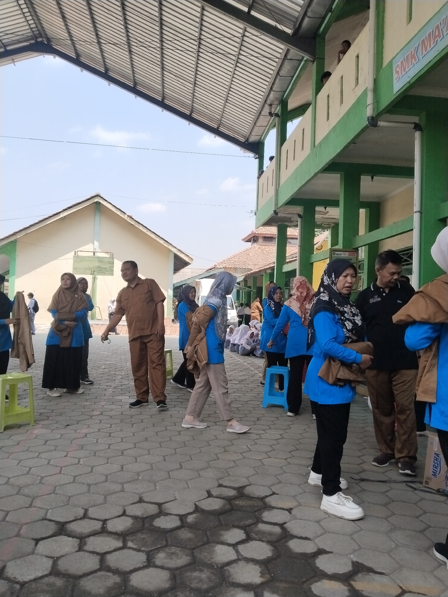 In Pakulaut, Indonesia, a group of individuals, predominantly women in blue shirts and headscarves, are gathered outdoors under a covered walkway. A man in a brown shirt and pants stands in the foreground, gesturing with his right hand. Another man in a black polo shirt is also present. The ground is paved with hexagonal concrete tiles. In the background, a two-story building with green and cream accents is visible, bearing the sign "SMK MAYA". Several people are seated in the background, and some are carrying jackets. The scene appears to be a casual gathering or event at a school.