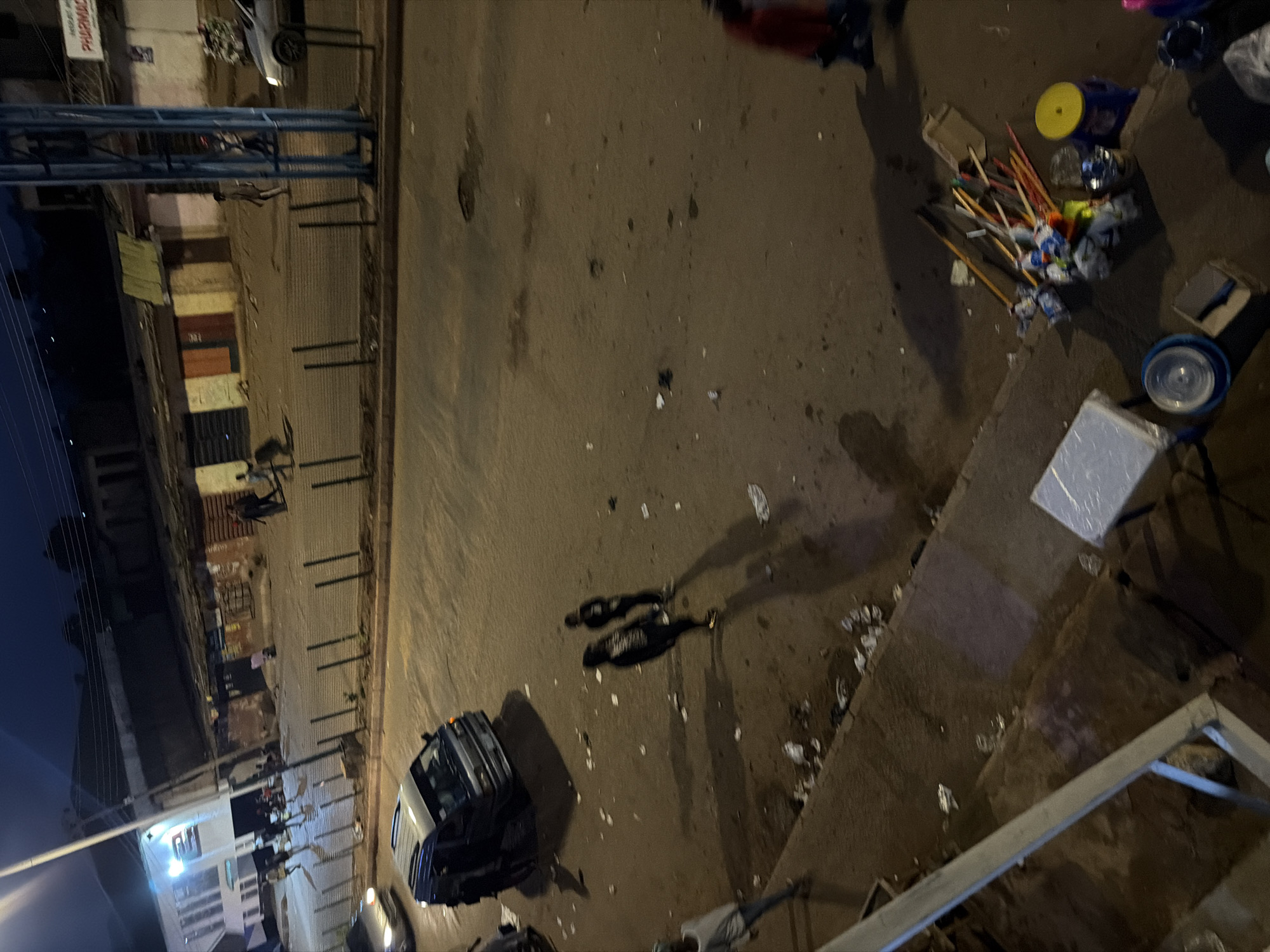 The image depicts a street scene at dusk in Bukuru, Nigeria. The street is paved with asphalt and shows signs of debris and litter scattered across its surface. On the left side of the street, there is a building with a brightly lit facade, possibly a shop or commercial establishment, with a prominent sign containing white lettering. Adjacent to this building, several people are walking or standing.
Further down the street, on the right, a row of small shops or market stalls with corrugated metal roofs are visible. A blue and white van is parked in the middle of the street, and two individuals are standing near it. In the foreground, two people are walking away from the camera, casting long shadows on the ground. To the right of the van, more people are visible, including one person wearing a red garment and another in blue.
In the lower right portion of the image, a collection of brooms, mops, and other cleaning tools are bundled together, along with a yellow-lidded container and several plastic buckets. A grey metal railing runs along the bottom left edge of the frame, suggesting a raised walkway or elevated area. The overall impression is of a bustling urban environment with ongoing activity, despite the dim lighting conditions.