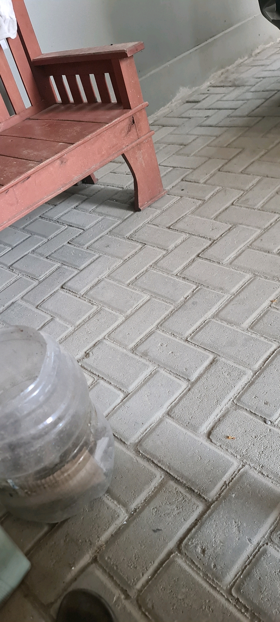 A close-up, eye-level shot shows a red wooden chair on a grey paver stone patio. The chair is weathered and appears to be an Adirondack-style chair, with a slatted backrest and armrests. A white cloth is draped over the left armrest. To the left of the chair, a clear plastic bottle is partially visible in the foreground. The patio pavers are laid in a herringbone pattern and are dusted with a light layer of debris. In the background, a grey wall forms a backdrop, and a portion of a dark vehicle is visible on the right side of the image. The scene suggests an outdoor setting, possibly a porch or patio area.