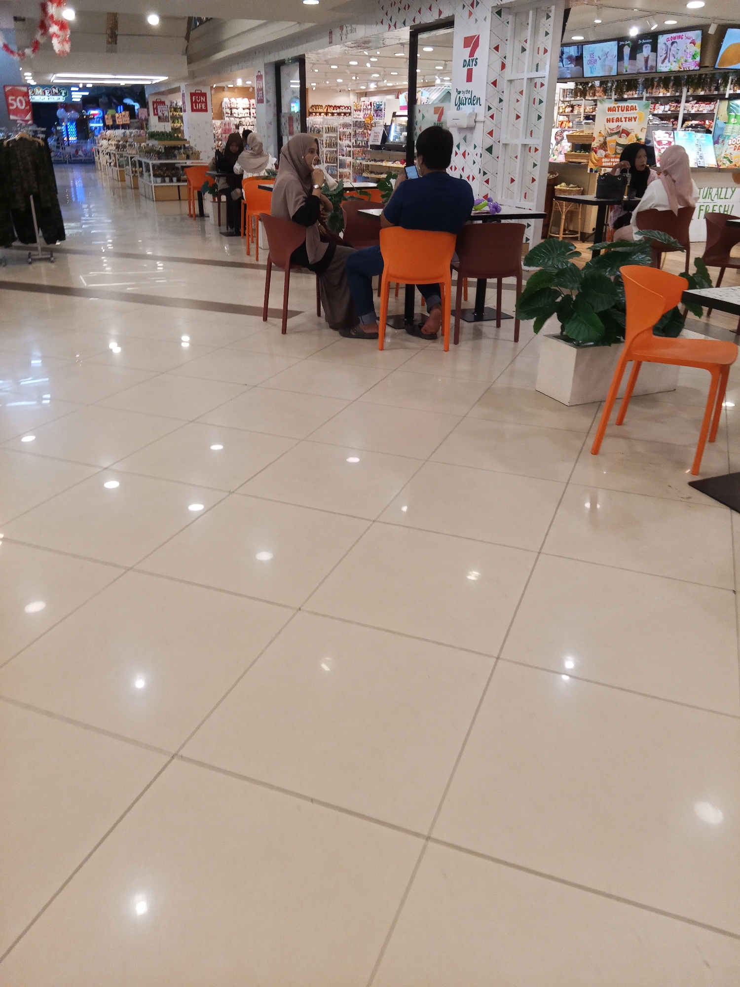 The image depicts an indoor scene, likely a shopping mall food court, in Lubuklinggau, Indonesia. Several individuals are seated at tables. In the foreground, a person in a blue shirt is looking at their phone, seated at an orange chair. To their right, a woman wearing a beige hijab is seated. Further back, two other women, one in a black hijab and another in a light pink hijab, are also seated at tables. The environment features tiled flooring with reflections from overhead lights, tables with chairs, and some potted plants. In the background, shop displays and promotional signage are visible. The overall atmosphere suggests a casual dining or resting area within a commercial space.