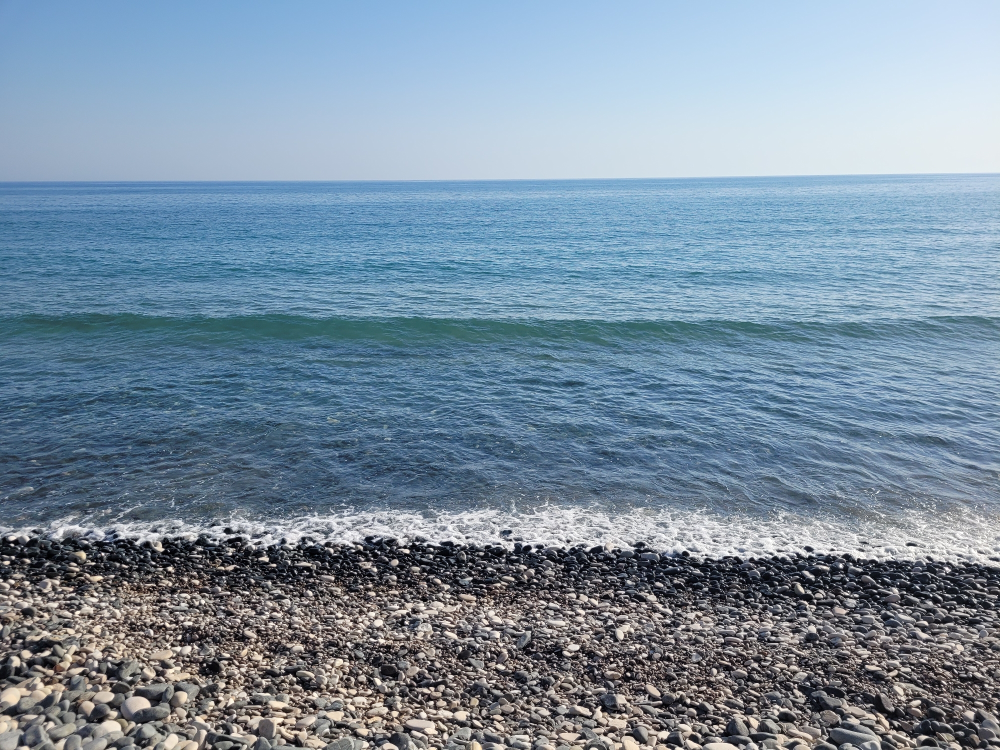 A calm, serene seascape is depicted, featuring a pebble beach in the foreground transitioning to the blue waters of the sea, which stretches to the horizon under a clear, pale blue sky. The pebbles on the beach are varied in size and color, ranging from light gray and white to dark gray and black, with a thin line of white foam from a gentle wave lapping at the shore. The water exhibits subtle ripples and variations in its blue hue, becoming a deeper, richer blue as it recedes towards the horizon. The scene appears to be a natural, undisturbed coastal environment, likely in Episkopi, Cyprus, given the context. There are no people or man-made objects visible in the frame, emphasizing the natural beauty of the location.