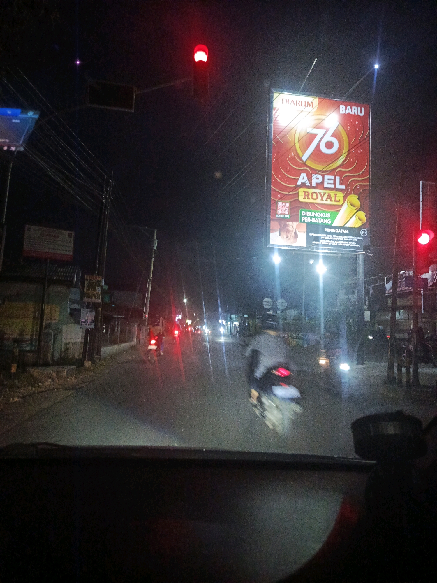 The image shows a nighttime view from inside a vehicle looking out at a street in Pejagoan, Indonesia. A large billboard advertising "DIARUM 76 APEL ROYAL" is prominently displayed on the right side of the street, illuminated by spotlights. A traffic light with a red signal is visible near the billboard. The street is paved and appears to be wet, reflecting the lights. Several motorcycles are present; one is in the foreground, blurred due to motion, and others are further down the road. Buildings with signs are visible on the left side of the street, along with power lines and streetlights. The overall scene depicts a typical urban street at night with ongoing traffic.