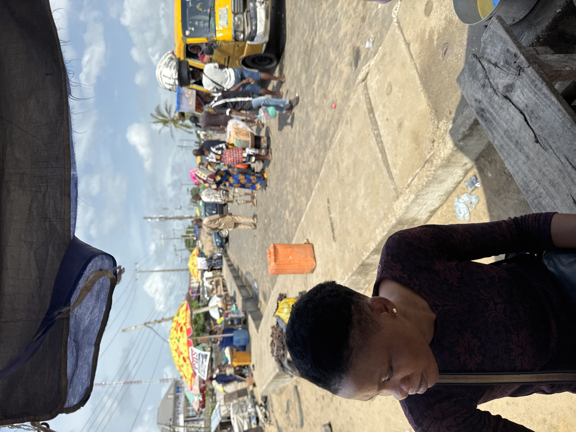 The image captures a street scene in Badagry, Nigeria. Focus is on a person with short dark hair, wearing a patterned long-sleeved top, seated near a weathered wooden surface. In the background, there is a bustling street with a yellow bus, several people walking, and vendors with colorful umbrellas and displays. There is an orange container near the center. The environment includes a mix of buildings, power lines, and a partly cloudy sky.