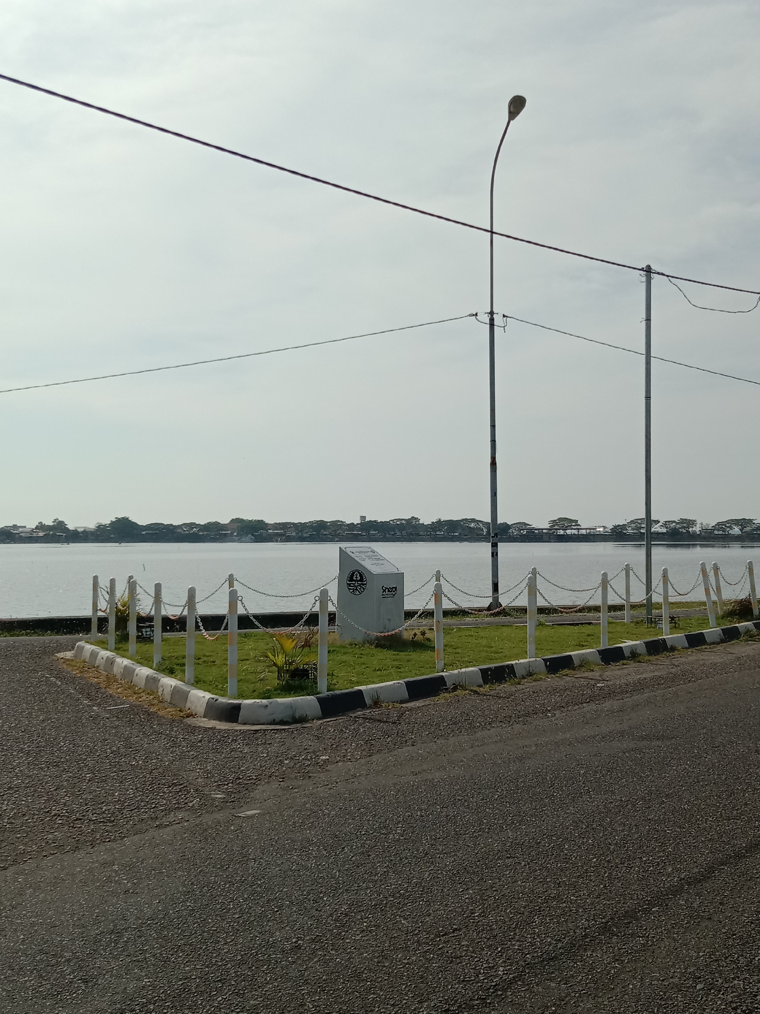The image presents an outdoor scene featuring a tranquil body of water under a bright, hazy sky. In the immediate foreground, a paved asphalt road curves slightly to the right, bordered by a curb painted with alternating black and white stripes. Adjacent to the curb is a small, well-maintained patch of green grass. This grassy area is enclosed by a low decorative fence composed of white posts connected by chains.
On the grassy section, a prominent white rectangular monument or sign stands, featuring a circular logo and the partially visible text "Senadi" beneath it.
Beyond the grassy area and fence lies a wide, calm expanse of water, likely a lake or reservoir, stretching across the middle ground. The water's surface subtly reflects the bright sky. Along the distant horizon, a strip of land is visible, dotted with numerous green trees and some scattered buildings, suggesting a coastal or lakeside settlement.
Overhead, several utility wires are stretched across the frame, supported by two tall utility poles on the right side. On the left, a street light with a curved pole extends upwards, positioned behind one of the utility lines.
The scene depicts daytime with soft, diffuse lighting, indicative of an overcast or hazy sky. There are no people visible, and no specific activities are taking place, creating a quiet and still atmosphere. The general setting, with the large body of water, road, and infrastructure, aligns with a waterfront area in Lhokseumawe, Indonesia.