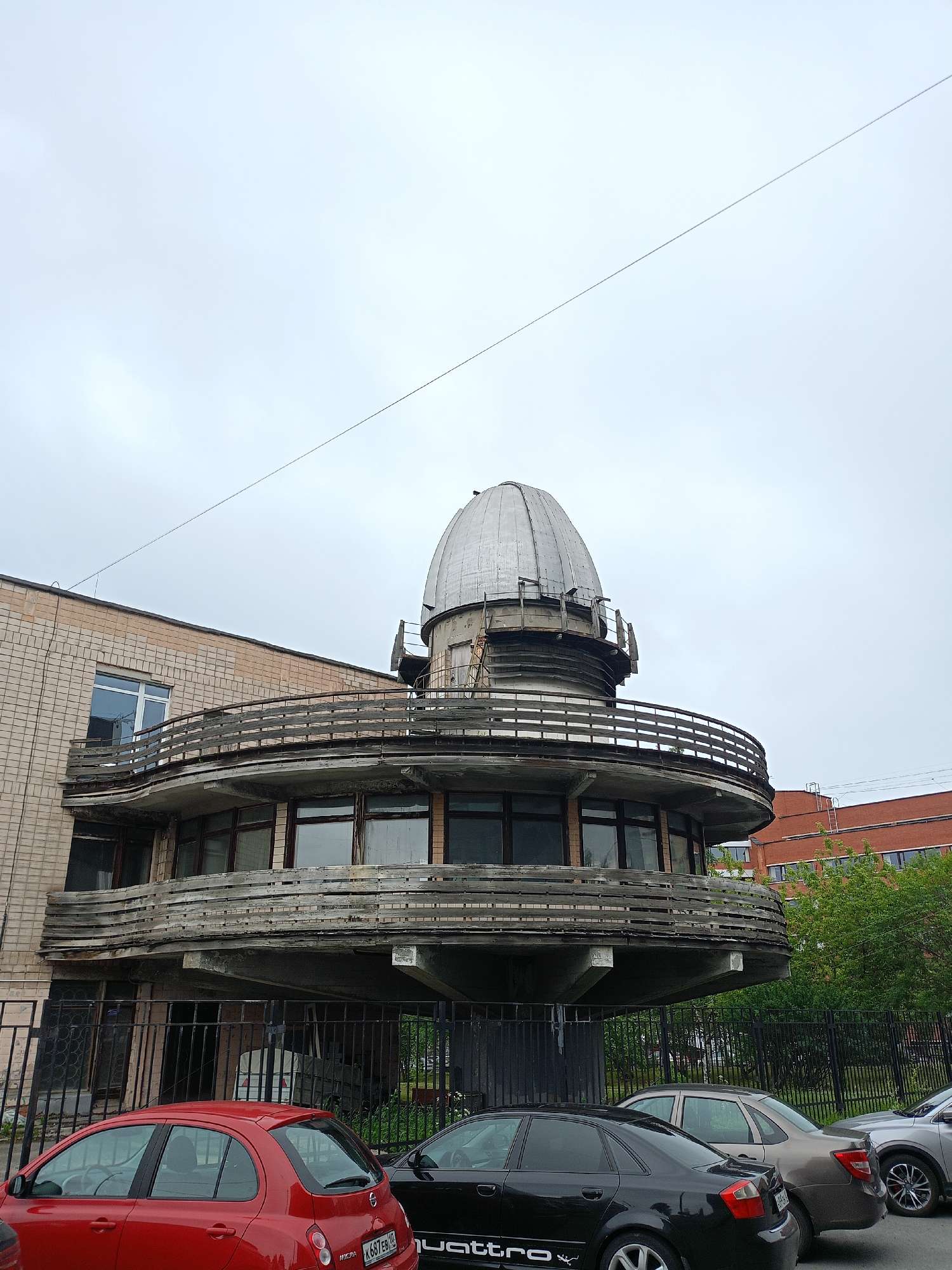This image captures an exterior view of a building with an observatory dome on top. The building itself appears to be made of light-colored brick, with a prominent, curved wooden balcony or walkway encircling a section of the structure. This balcony has a weathered, aged appearance. Below the balcony, there are large windows.
In front of the building, there is a black metal fence. Beyond the fence, several cars are parked, including a red hatchback, a black sedan, a beige sedan, and a silver SUV. The sky is overcast, suggesting a cloudy or perhaps rainy day. The lighting is diffused and even, indicating it is likely daytime.
To the right of the main building, a taller, reddish-brown brick building is visible, with multiple windows and a visible fire escape or ladder structure. Trees and greenery are also present behind the fence and around the buildings. A thin wire stretches diagonally across the upper portion of the image against the sky.
No people are visible in the image. The scene appears to be a quiet, possibly institutional or research facility, given the observatory dome. The overall impression is one of functional architecture, with some elements showing signs of age and weathering.
Visible text includes a license plate on the red car, which appears to be "K687EB." On the black sedan, the word "quattro" is visible.
