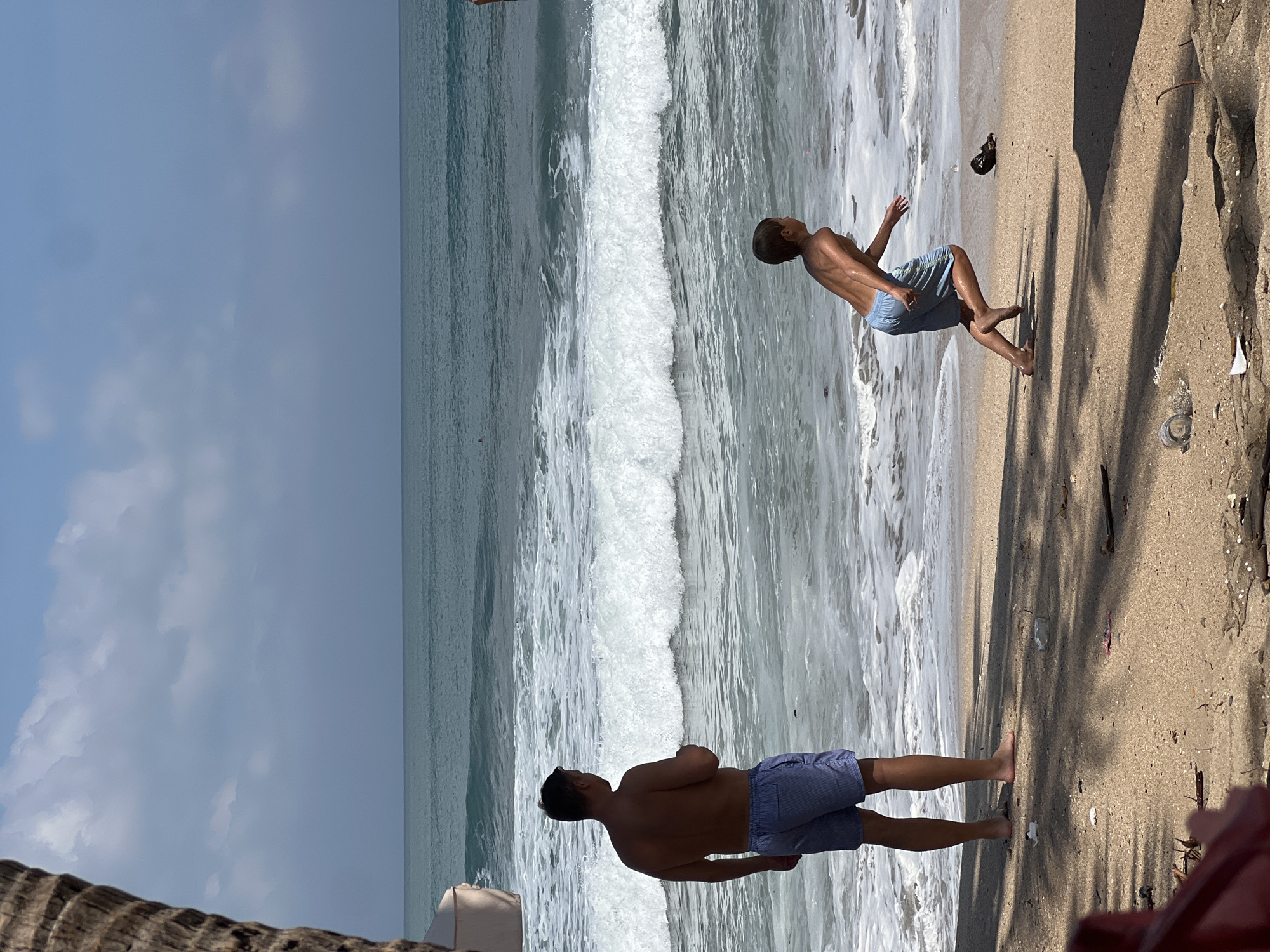 An adult male stands on the sandy shore of Kuta Beach, Indonesia, facing the ocean. He is wearing blue patterned swim shorts. To his right, a young boy, also in blue swim shorts, runs towards the water's edge. The ocean has moderate waves breaking on the shore, creating white foam. The sky is partly cloudy, with a blue hue. The foreground shows the sandy beach with scattered debris, including a plastic bottle. A palm tree trunk is visible on the far left, casting a shadow on the sand.