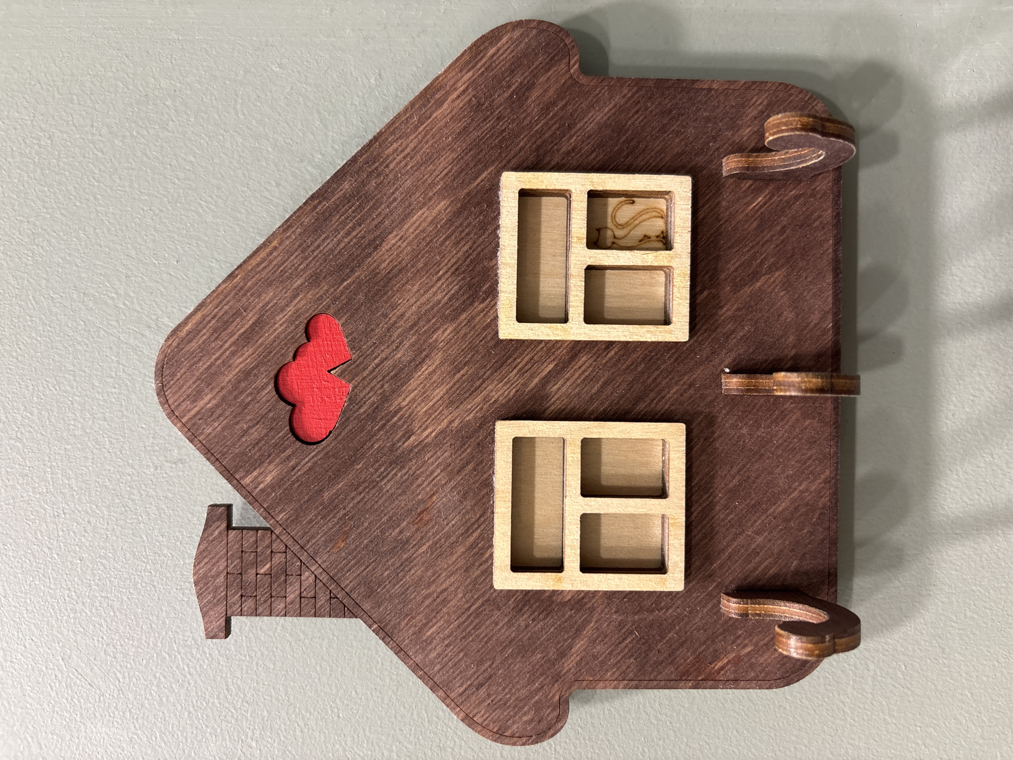 The image displays a stylized, house-shaped wooden wall decoration mounted against a textured, light gray wall. The main structure of the house is crafted from dark brown wood, featuring a visible grain and diagonal etched lines that add texture.
Two light-colored, rectangular wooden window frames are positioned vertically on the right side of the house, one above the other. Each window contains internal dividers, creating multiple panes. Within the upper right pane of the top window, a small, intricate cutout of a stylized bird, resembling a swan or goose, is embedded.
On the left side of the house, beneath the roof peak, a vibrant red, irregularly shaped wooden piece, reminiscent of a cloud or a simplified bush, is set into the dark brown wood. At the bottom left, a lighter brown wooden piece with etched lines suggesting bricks or shingles protrudes, forming a detail that resembles a chimney or a small architectural extension.
Along the right edge of the house, three small, curved, layered wooden elements are attached. These pieces exhibit alternating light and dark wood layers, indicating they are composed of multiple thinner pieces, and function as decorative accents or possibly hooks.
The item appears to be a handcrafted or laser-cut decorative piece, characterized by its clean lines and contrasting wood tones. No people are present, and there is no visible text. The lighting suggests an indoor setting.
