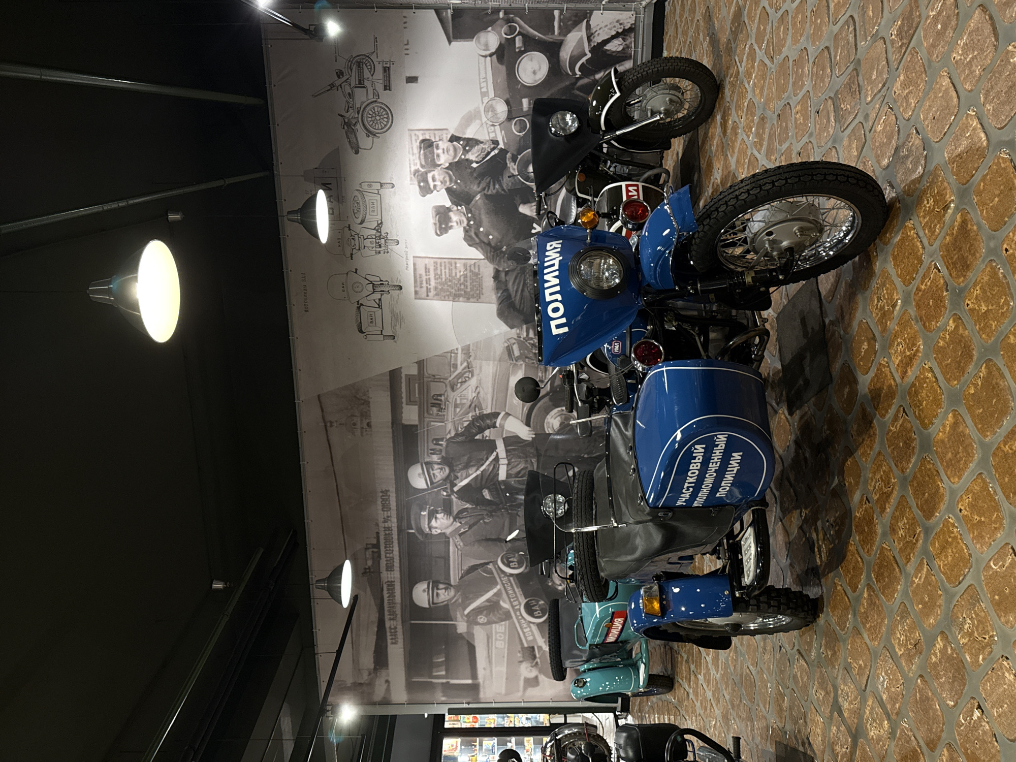 In the city of Arkhangel’skoye, Russia, this image depicts a museum interior. The foreground is dominated by two blue motorcycles with sidecars. The motorcycle on the right is black, and the one on the left is turquoise. Both are parked on a floor covered with rectangular stone tiles. A large mural covers the wall behind the motorcycles, featuring vintage black and white photographs of police officers and schematics of motorcycles with sidecars. The ceiling is dark, with industrial lighting fixtures.