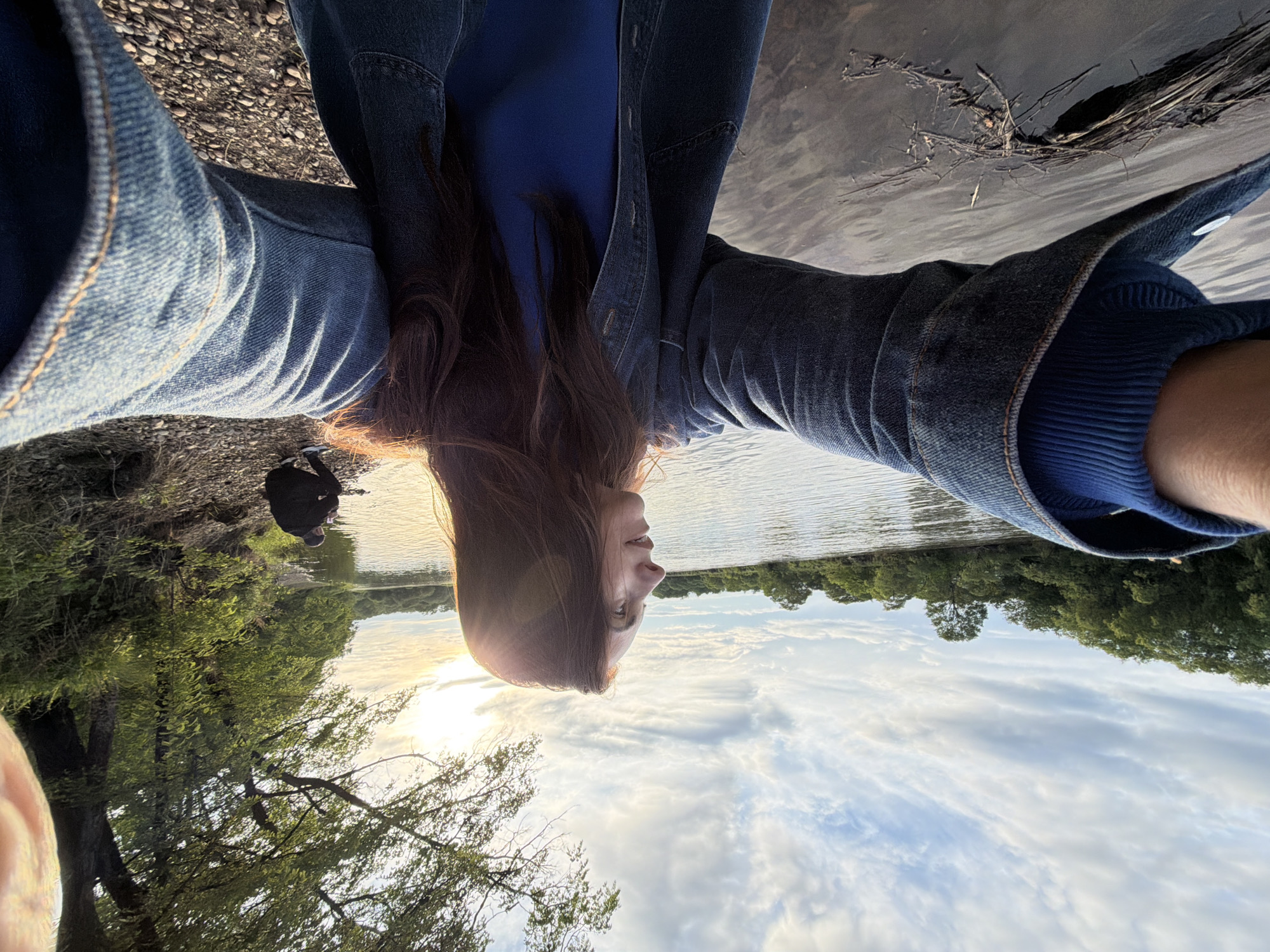 The image displays an outdoor scene by a river in Birobidzhan, Russia, during what appears to be the golden hour, characterized by warm, low-angle sunlight. In the foreground, a female with long, dark brown hair, wearing a blue denim jacket over a blue ribbed long-sleeve top, takes a wide-angle selfie. Her head is turned in profile to the right, looking towards the sun, which creates a strong lens flare and backlighting effect around her head and hair. Her arms are outstretched, holding the camera.
The woman stands near the left bank of a dark, calm river or large stream, which occupies the lower-left and central portions of the image. Floating debris, including sticks and branches, is visible in the water near the foreground bank. The opposite bank (right side of the frame) features a rocky shoreline leading to a dense line of green deciduous trees and bushes. Further along this right bank, a second individual, dressed in dark clothing, is crouched at the water's edge. The sky above is partly cloudy, with scattered stratocumulus and wispy cirrus clouds illuminated by the setting or rising sun on the right. A large tree branch with green leaves extends into the upper right corner of the frame.