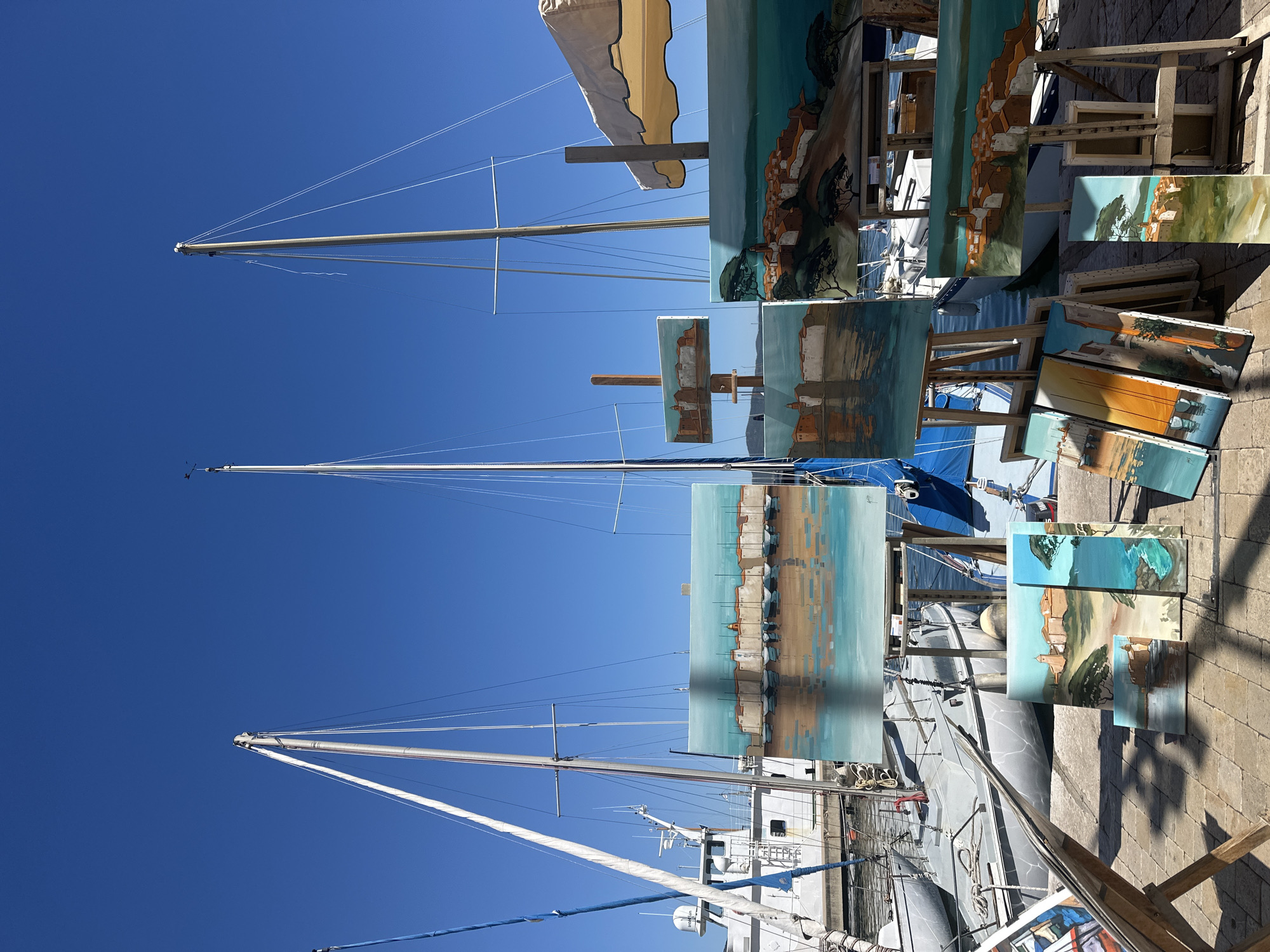 An outdoor art display of several paintings is set up on a paved walkway in a harbor. In the background, several sailboats are docked, and a larger white boat is visible on the left. The sky is clear and blue. The paintings are of coastal scenes, featuring buildings, water, and boats, with some depicting reflections. The art display appears to be situated along the waterfront in Saint-Tropez, France. No people are visible in the image.