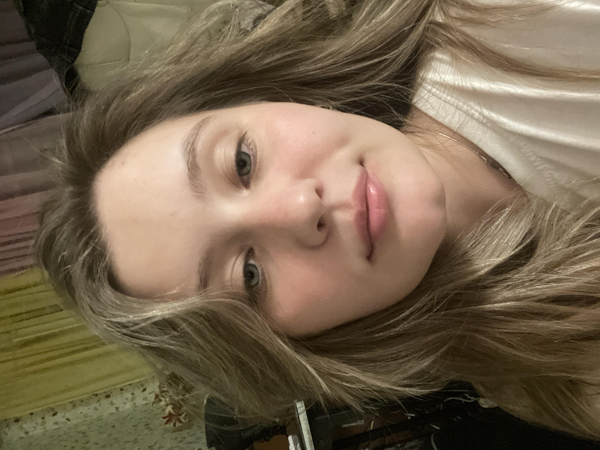 A close-up, head-and-shoulders portrait shows a young woman with long, blonde, wavy hair and fair skin. Her blue eyes are looking slightly to the left, and her lips are slightly parted in a faint smile. She is wearing a white t-shirt, and a delicate silver necklace is visible around her neck. The background is softly blurred, with hints of yellow and purple fabric suggesting curtains or drapery. There are also dark shapes that could be furniture or other objects in the room. The lighting is dim, creating a warm, intimate atmosphere.