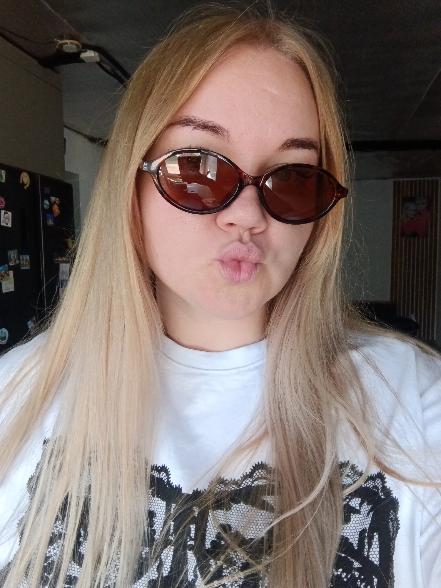 A blonde-haired woman with fair skin wears brown oval sunglasses and a white t-shirt with a black lace-like design. She is pouting her lips towards the camera. The background includes a refrigerator with magnets and photos, a ceiling fixture, and a partially visible framed photo on a wall. The location in the city of Фоки, Russia is not discernible from the image.