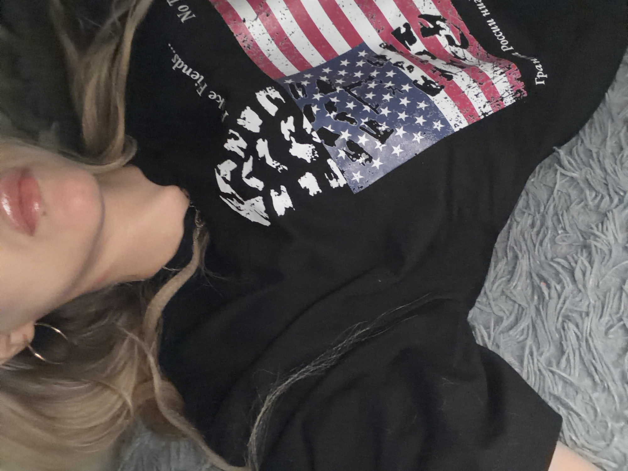 A close-up, overhead shot shows a person lying on a gray fuzzy blanket. The person has blonde hair and is wearing a black t-shirt with an American flag graphic and text. The text on the shirt is partially visible and appears to be in English and possibly Russian. The person is wearing a hoop earring. The image focuses on the upper chest area and face, which is partially obscured.