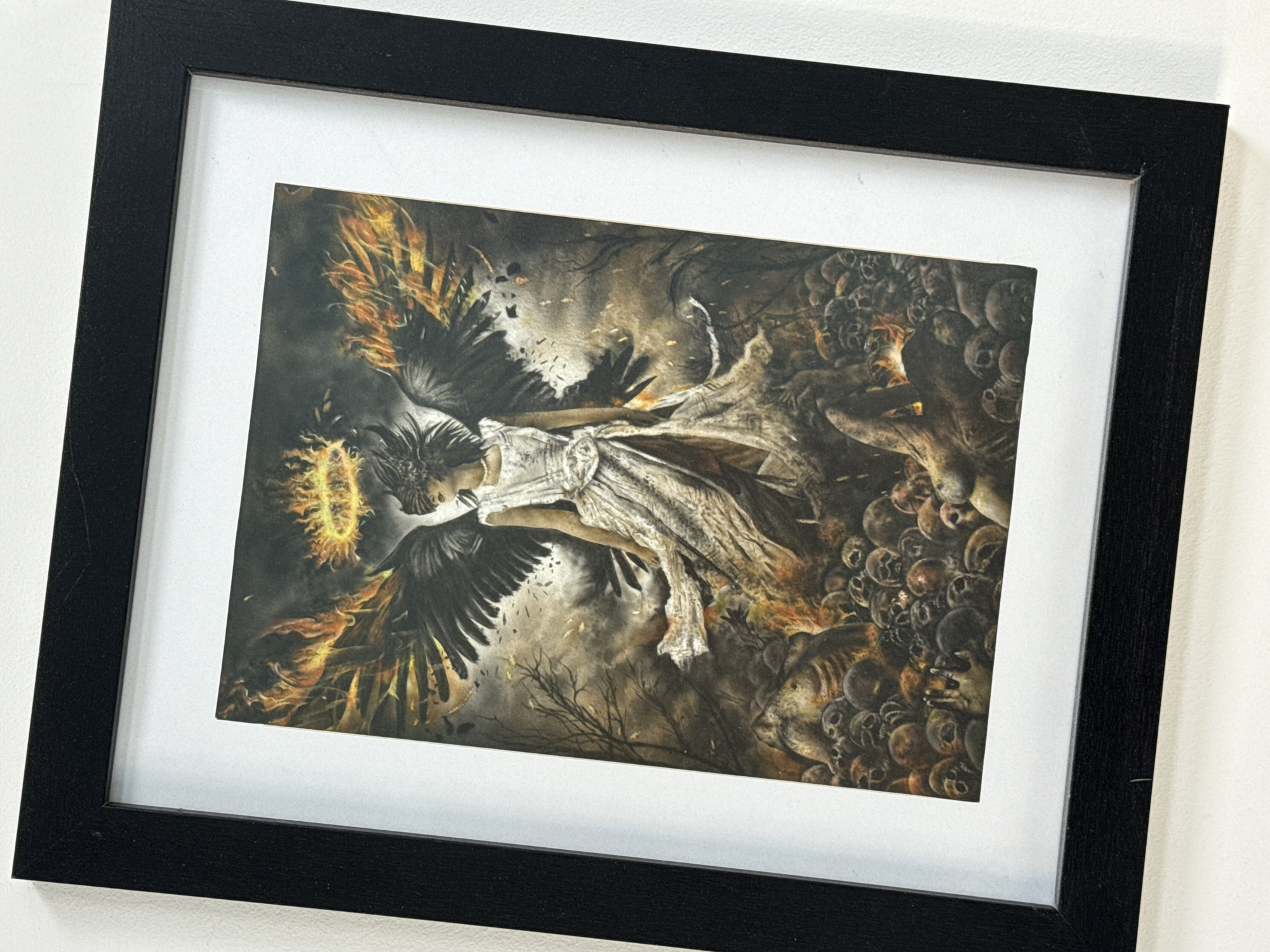 A framed print of an elaborate, dark fantasy-themed artwork hangs on a white wall. The print features a figure that seems to be an angel or a similar being with dark wings, surrounded by flames and skulls. The figure is partially adorned in what appears to be a white dress, and the surrounding environment is dark and complex, featuring skeletal elements and possibly trees. The frame is black and rectangular, and it is set against a plain white wall. There is no contextual evidence to indicate a location in Burgas, Bulgaria.