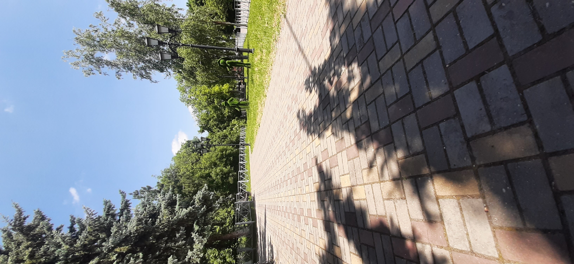 This image depicts a park scene in Yessentukskaya, Russia. The viewpoint is low, looking up at a bright blue sky with scattered white clouds. On the left, a row of large evergreen trees with distinctive blue-green needles stands prominently. Beyond them, lush green deciduous trees form a dense backdrop.
A paved walkway, made of reddish-brown and gray rectangular bricks laid in a herringbone pattern, dominates the right side of the frame and recedes into the distance. The walkway is partially covered by dappled shadows, suggesting a sunny day with trees casting their silhouettes.
Along the walkway, a white metal fence runs parallel, separating the path from a neatly mown green lawn. Several black lampposts with multiple fixtures are visible on the lawn, spaced along the fence. In the background, two topiary figures, appearing to be shaped like people and covered in green material, stand near one of the lampposts. The overall impression is one of a well-maintained park on a pleasant day. The time of day appears to be daytime, given the strong shadows. There is no visible text in the image.