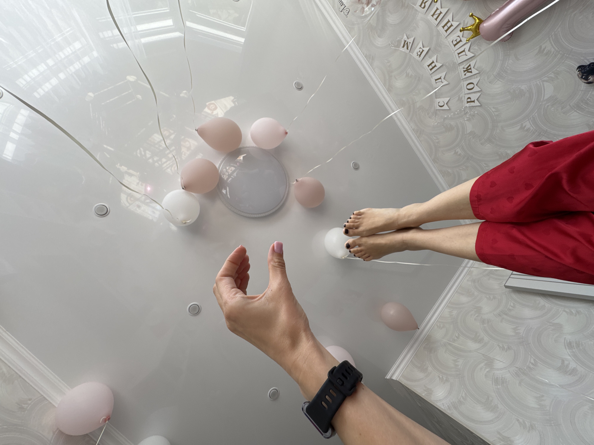 A low-angle shot captures a person's hand and arm, adorned with a black smartwatch, extending towards a white ceiling. Scattered across the ceiling are several light pink and white balloons tethered with ribbons. A person's legs and feet are also visible, wearing red shorts. The background features a decorated wall with celebratory lettering, possibly in Russian, and decorative crown imagery, suggesting a birthday theme. The room has a modern, well-lit interior. There is no visible evidence to determine the specific location in the city of Sochi, Russia.