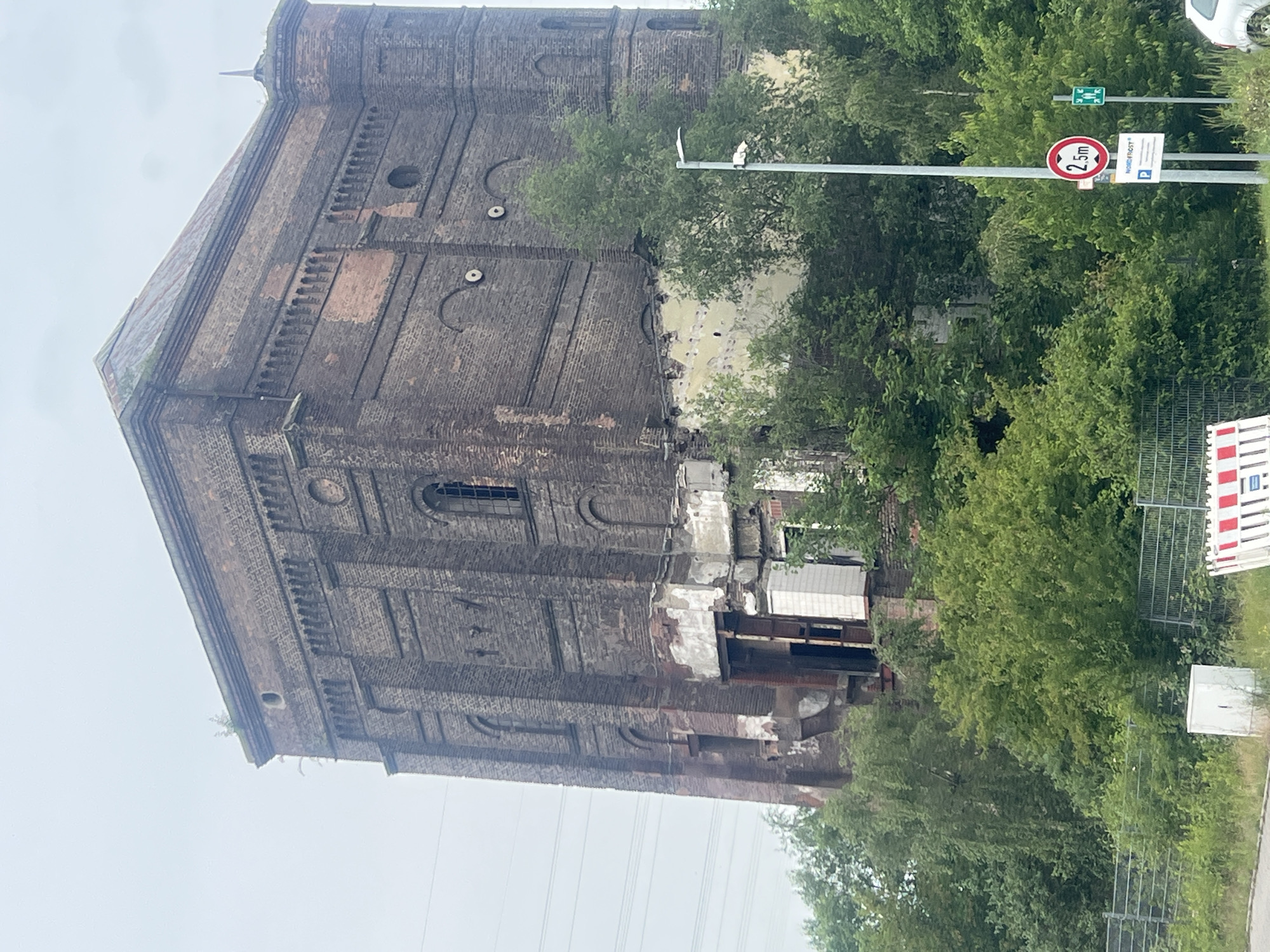 The image shows a tall, derelict brick building with a pyramidal roof. The building appears to be an old industrial structure, possibly a water tower, given its shape and the surrounding industrial landscape. The brickwork is dark and weathered, with sections of it crumbling and exposing the inner structure. There are several arched windows, some of which are broken or boarded up. The building is surrounded by dense green foliage, with trees growing up around its base and obscuring some of its lower sections. In the foreground, there is a metal fence, a white rectangular box, and a traffic barrier with red and white stripes. A lamppost stands near the fence, with a red circular sign showing "2.5m" and a green rectangular sign. The sky is overcast. The building is located in the industrial area of Herne, Germany.