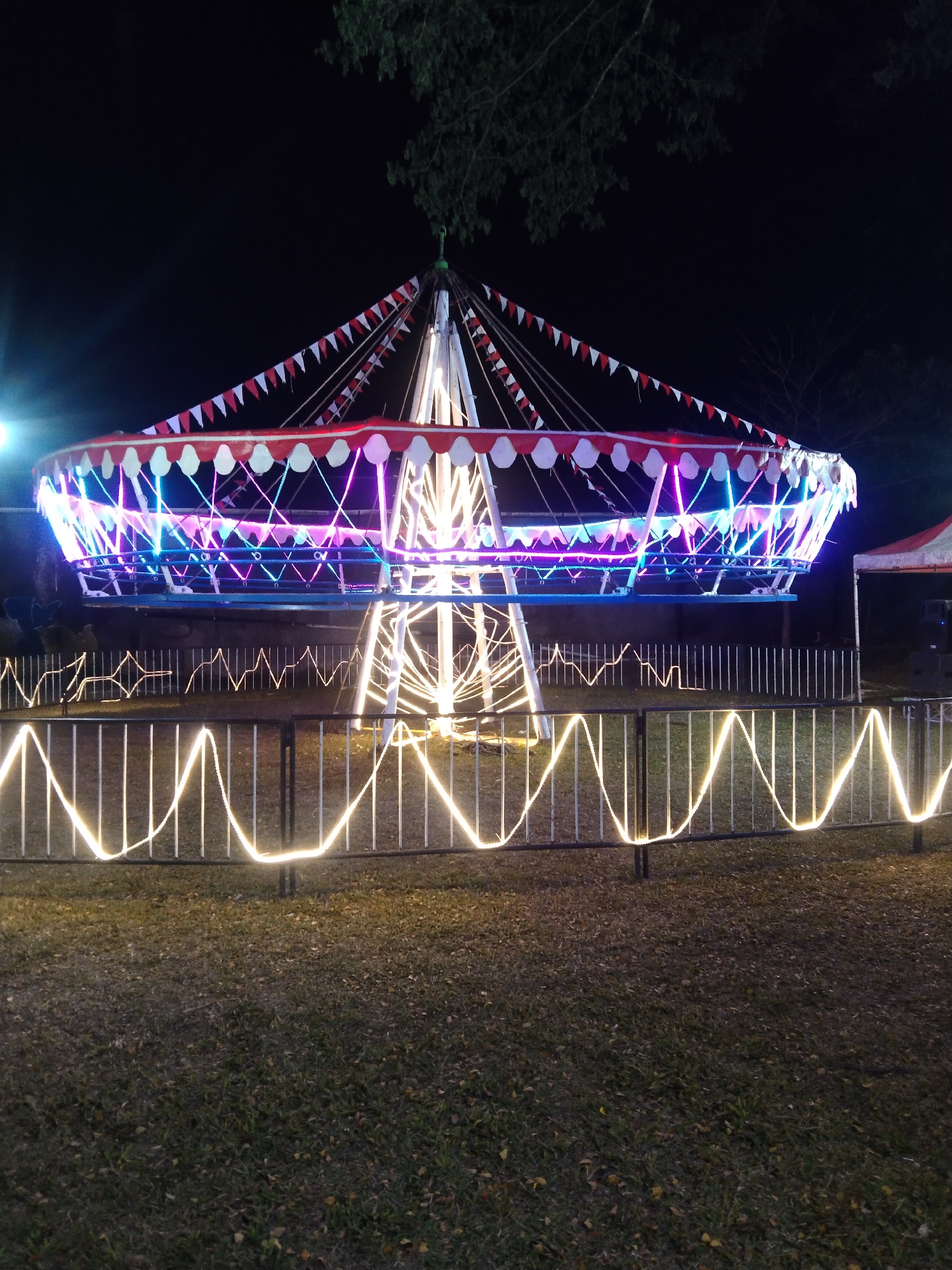 The image depicts a brightly lit carousel at night, likely set up in an open area such as a park or field. The carousel is illuminated with colorful LED lights, predominantly blue and pink, creating a vibrant spectacle against the dark night sky. Red and white pennant flags adorn the top of the carousel structure, adding a festive touch. A metal fence with yellow string lights, forming a wave-like pattern, separates the viewer from the carousel. The ground in the foreground is covered in grass and scattered leaves. The background is dark, with hints of trees and foliage visible above the carousel. A red and white striped tent is partially visible on the right side of the image. This scene is likely from a public event or attraction in Magelang, Indonesia, given the request.