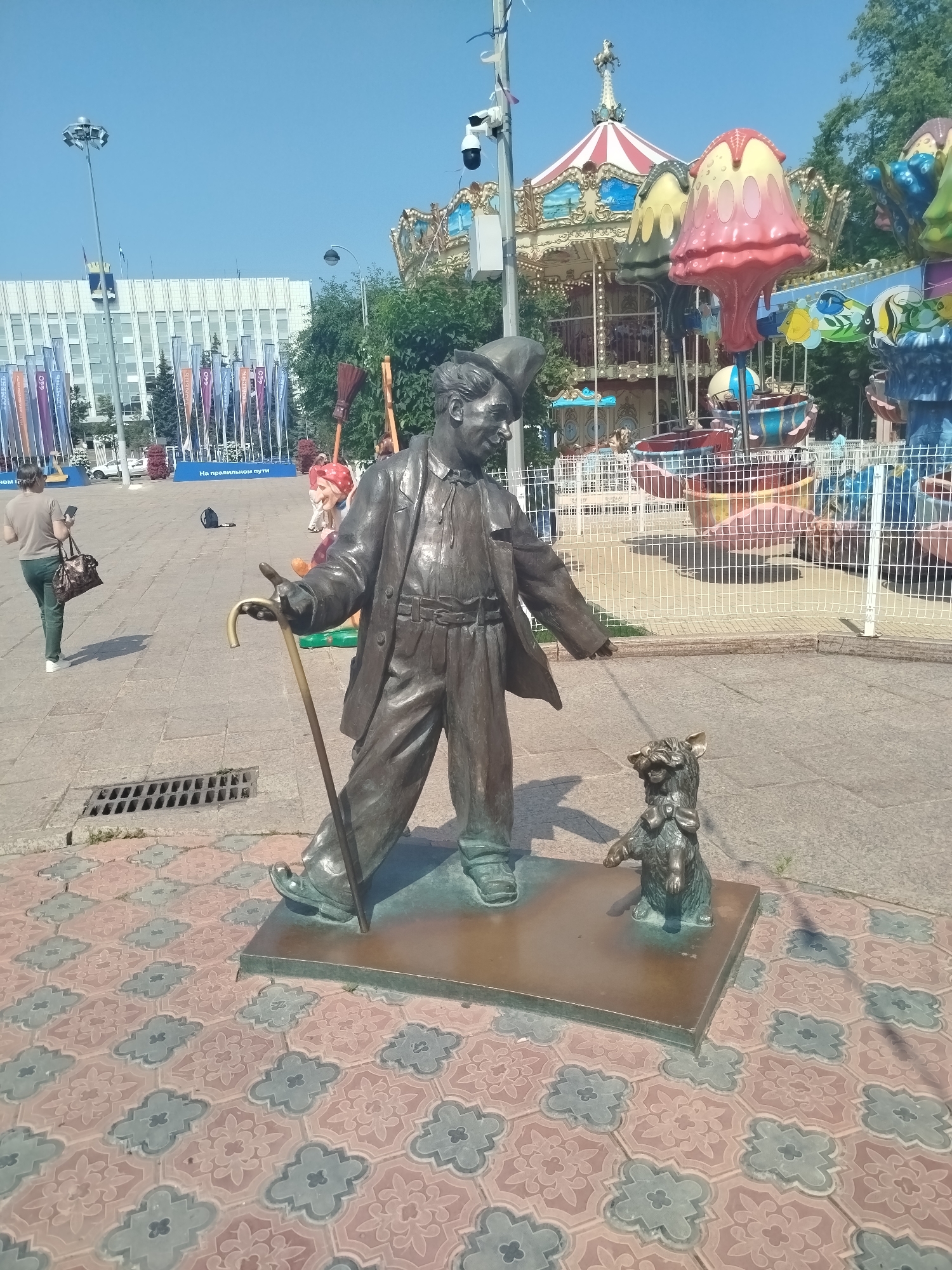 The image shows a bronze sculpture of a man and a dog on a patterned tiled ground in a city square. The man, wearing a hat and suit, is holding a cane and appears to be looking down at the dog, which is sitting with its head tilted. In the background, there is a carousel with colorful rides, a building with flags, and trees. A woman with a bag and phone walks away from the camera on the left. There is also a street drain grate visible in the foreground. The scene is set on a bright, sunny day with a clear blue sky. This sculpture is located in the Embankment of the Tura River in Tyumen, Russia, and depicts characters from a popular Soviet-era film.