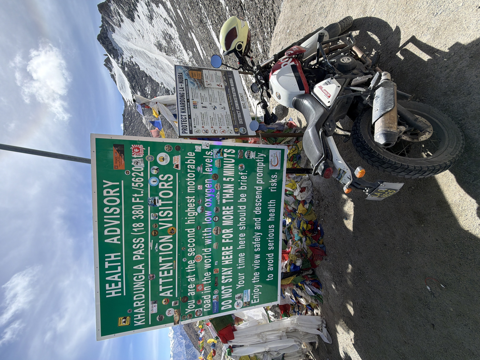 The image depicts the Khardungla Pass, a high mountain pass in the Ladakh region, near the city of Leh, India. A large green sign prominently displays a "HEALTH ADVISORY" for Khardungla Pass, stating it is at 18,380 ft (5620 m) and is the second highest motorable road in the world with low oxygen levels. It advises visitors not to stay for more than 5 minutes due to potential serious health risks from the low oxygen. The sign is adorned with numerous stickers, including travel-related logos and warnings.
To the right of the main sign, a smaller sign titled "PROTECT KHARDONG LA - NUBRA" provides further instructions on responsible tourism, such as "KEEP IT CLEAN" and "PRESERVE NATURE."
A white, red, and black motorcycle is parked on the dirt and gravel road in front of the signs. A yellow helmet with a smiley face design is placed on the handlebars. Prayer flags, a common sight in mountainous regions of India and Nepal, are draped around the base of the signs and on a nearby pole. The background features a steep, snow-capped mountain range under a partly cloudy sky. The ground is a mix of gravel and dirt, with shadows cast by the signs and the motorcycle.