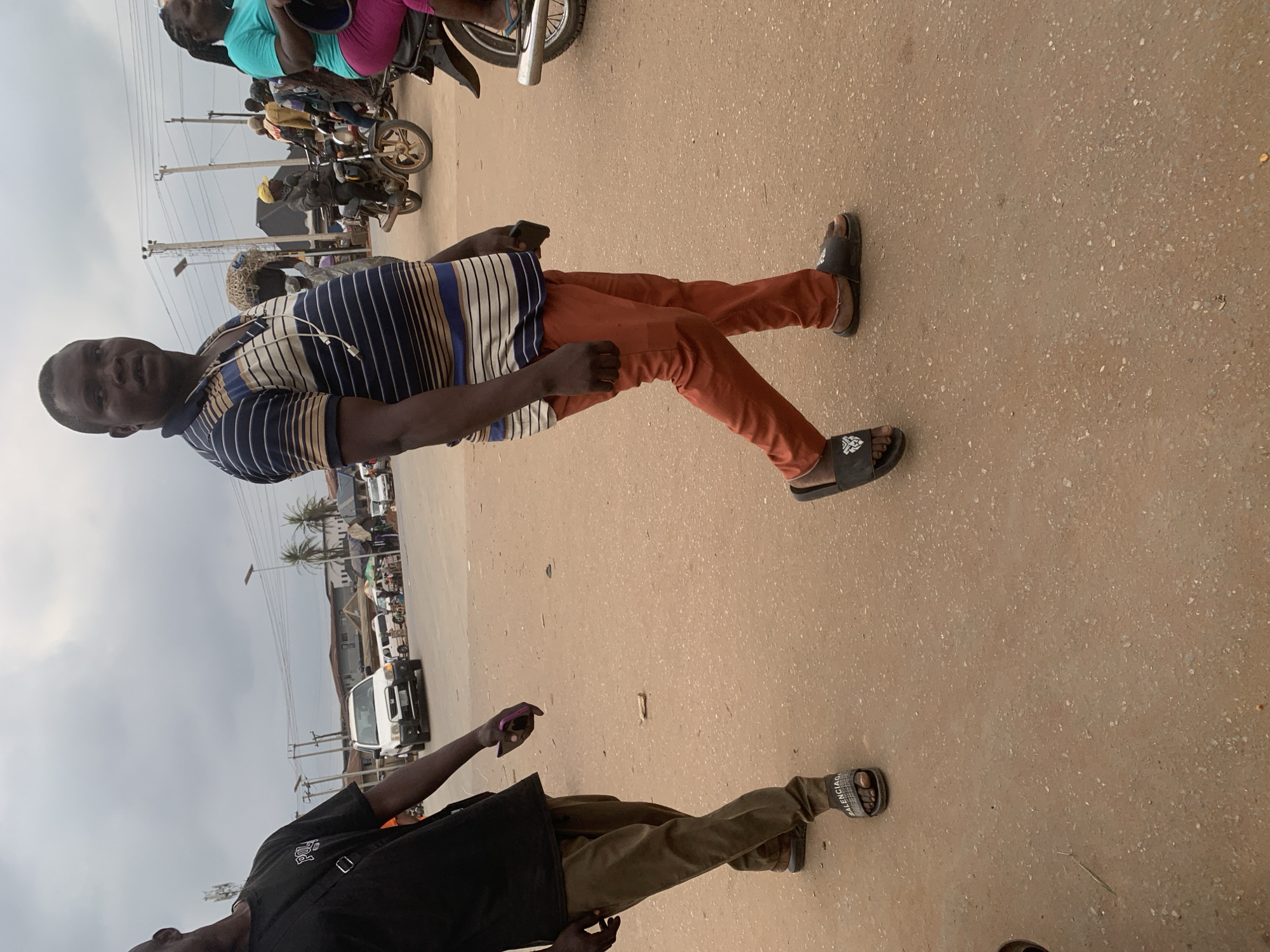 The image depicts a street scene in Shagamu, Nigeria, under an overcast sky. In the foreground, a Black man walks towards the right, wearing a horizontally striped polo shirt in blue, white, and brown, and orange pants. He has a dark-colored phone in his right hand. To his left, another Black man walks towards the left, wearing a black t-shirt and khaki pants, carrying a black bag over his shoulder and holding a purple object in his right hand. The ground is a dusty, sandy road. In the background, several motorcycles are visible, with people riding them and standing around. One motorcycle has a rider wearing a yellow cap. Further in the background, there are buildings, power lines, and some palm trees. A white vehicle, possibly an SUV, is parked on the left side of the road, and other vehicles are seen in the distance. The overall impression is a busy street environment.