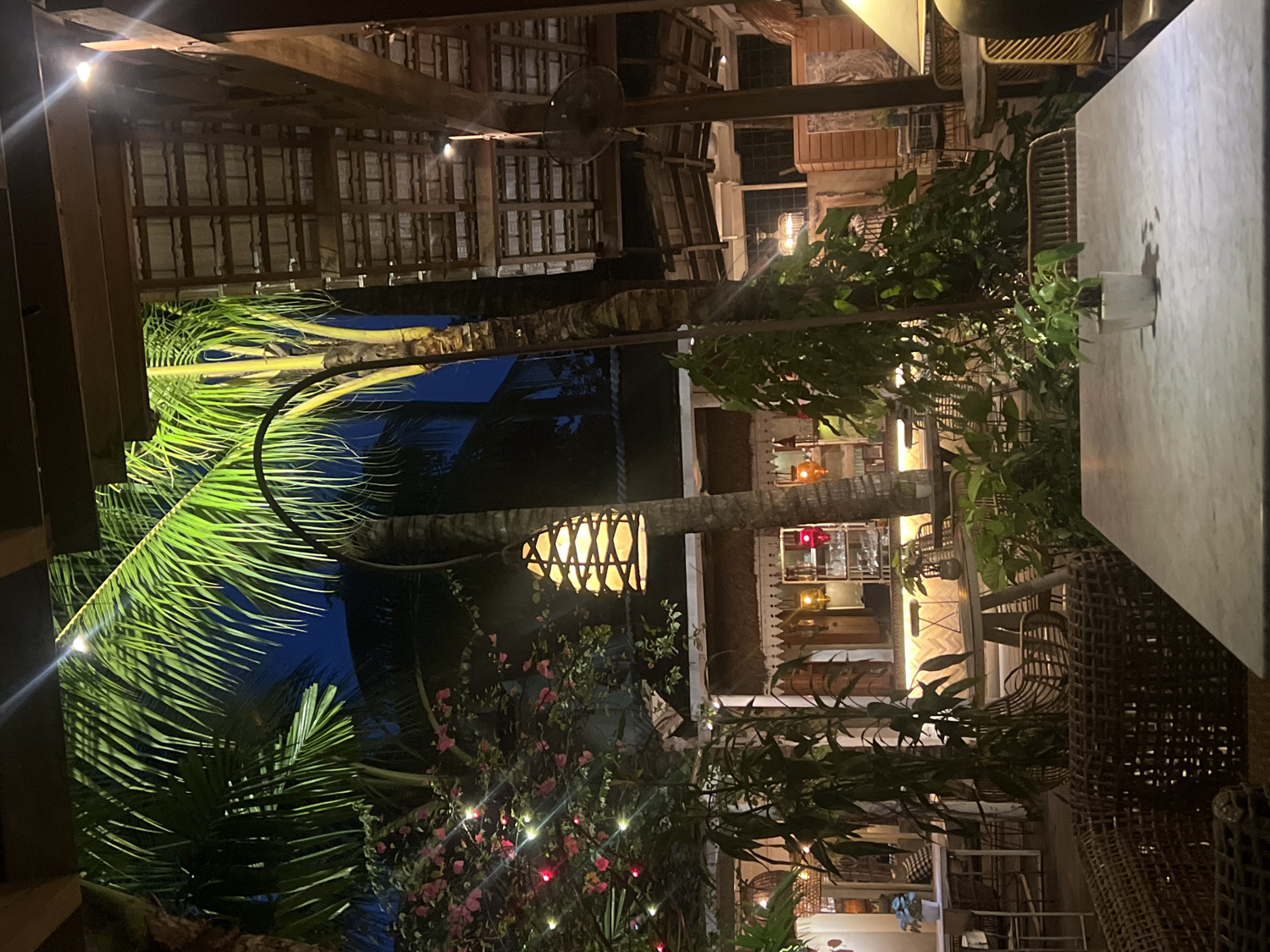 The image depicts an illuminated outdoor or semi-outdoor tropical setting at night, likely a dining or lounge area within a resort or restaurant in North Kuta, Indonesia. The scene is characterized by lush vegetation and warm artificial lighting, creating a relaxed and inviting atmosphere.
In the foreground, a light-colored, possibly marble or stone, table occupies the right side of the frame, hinting at a dining or social space. To the left and center, prominent palm trees feature expansive, bright green fronds that are dramatically lit, contrasting against the dark night sky. A distinctive woven lantern hangs from one palm tree trunk, emitting a soft, warm glow. Below, string lights with red bulbs are interspersed among smaller bushes and plants, adding a festive sparkle to the lower foliage.
Above, a rustic wooden ceiling structure with exposed beams and planks is visible, featuring overhead lights and a ceiling fan, suggesting a covered area. In the middle and background, a series of charming, traditional-style buildings are visible, characterized by their wooden details, gabled roofs, and warm interior lighting. These structures appear to house further seating areas, furnished with wicker or rattan chairs and tables.
No individuals are present in the image, contributing to a tranquil and peaceful ambiance, perhaps captured during a quiet moment. The overall impression is one of an exotic and comfortable evening retreat. There is no visible text in the image.
