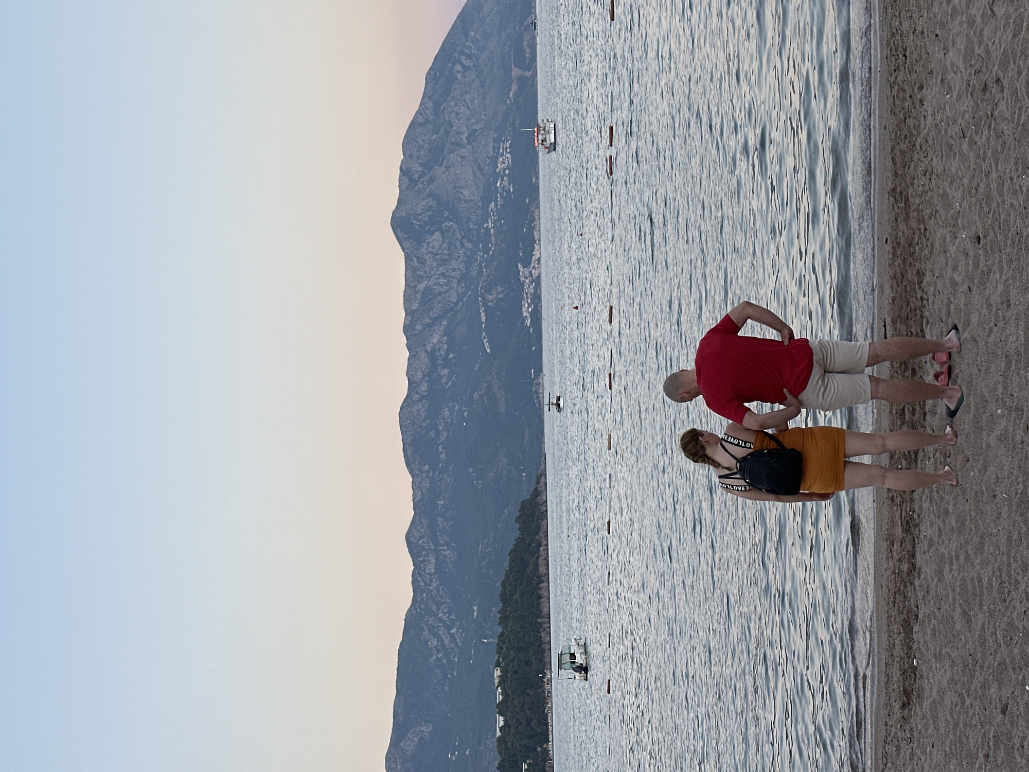 Two people, a woman and a man, stand on a sandy beach facing a calm sea with a mountainous coastline in the background. The woman is wearing a short yellow dress and a black backpack, with her hair in a braid. The man is wearing a red t-shirt and light-colored shorts. They appear to be looking out at the water. Several boats are visible on the sea, and a line of buoys stretches across the water in the middle ground. The sky is a pale gradient of blue and pink, suggesting it is either dawn or dusk. The scene is likely located in or near Budva, Montenegro, given the distinctive mountainous coastline.