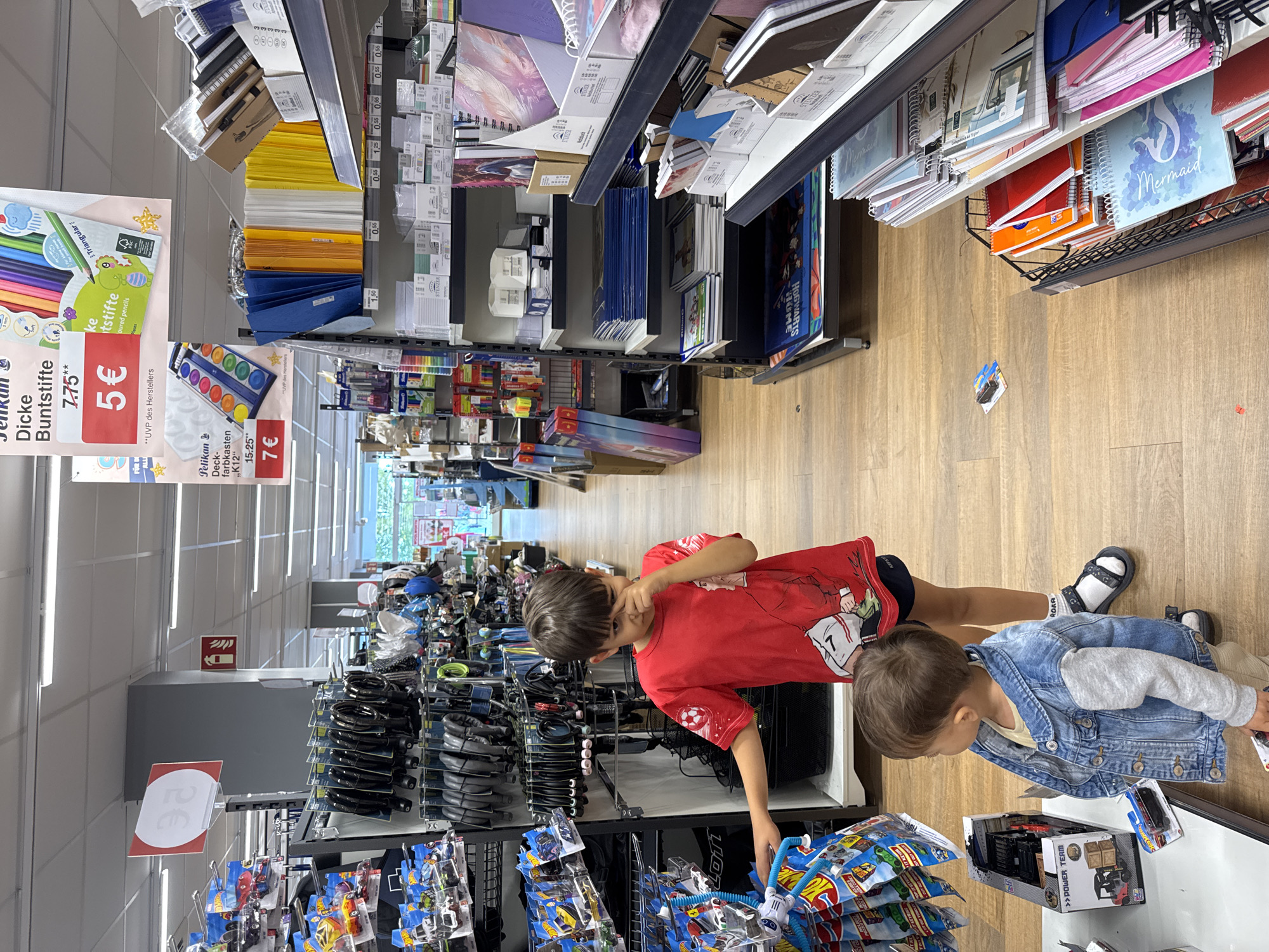 A young boy in a red t-shirt and shorts stands in an aisle of a store, with his younger brother in a denim jacket and beige pants beside him. The store appears to be a stationery or toy shop. The older boy is pointing to something on a shelf, and the younger boy is looking down at a toy car. The shelves are stocked with various items including toys, stationery, and art supplies. In the background, there are more shelves filled with merchandise, and the store appears to be well-lit with fluorescent lighting. The floor is made of light-colored wood. There are promotional signs visible on the shelves. The setting is likely a retail store in Saarlouis, Germany, given the text on the signs, which appears to be in German.