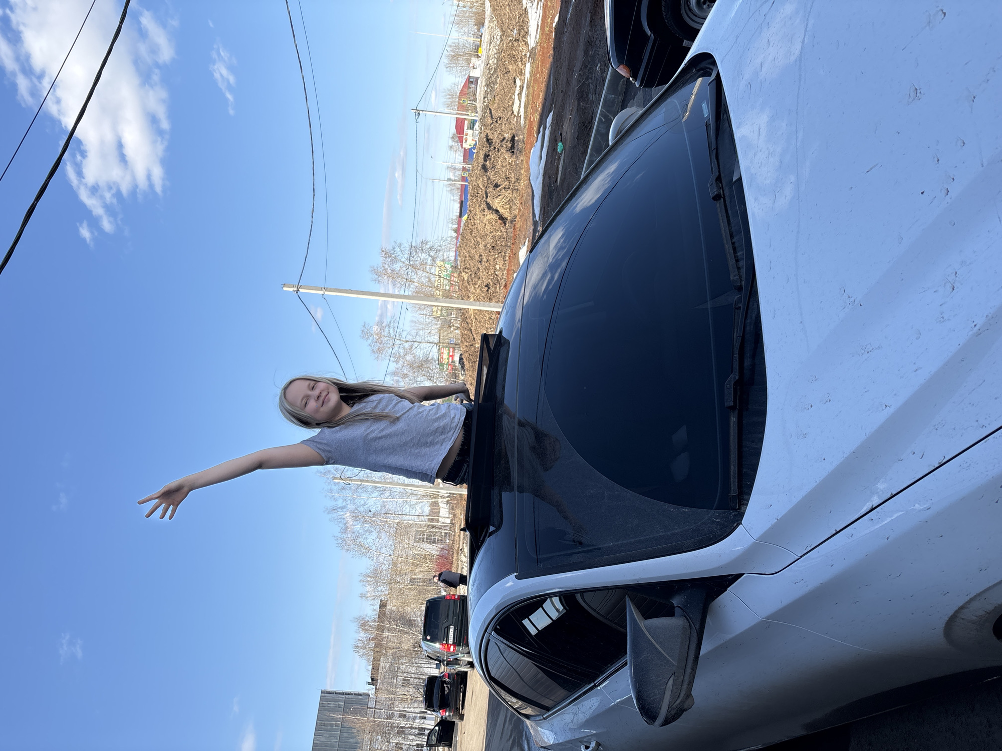 A girl is standing in the open roof of a white vehicle. She is extending her right arm outwards. The car is parked on a street in Izhevsk, Russia. Other vehicles are behind her. In the background, there is some construction and trees. The sky is blue with some clouds. Overhead power lines are visible.