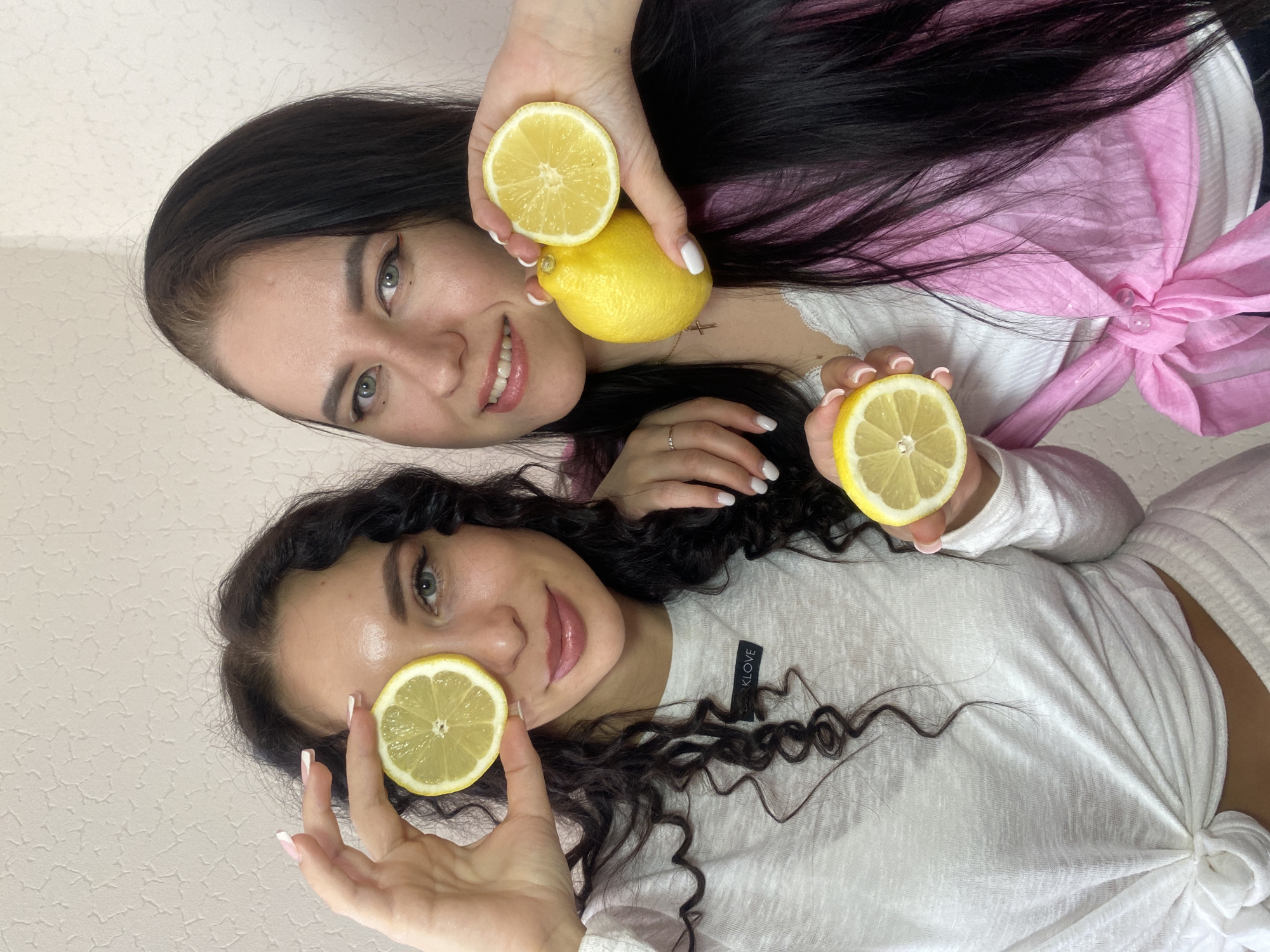 Two young women are cheerfully posing for a photo indoors, each holding lemons. The image is oriented vertically, appearing as if the subjects are lying down or the camera was held at an angle.
The woman on the left (as viewed when the image is upright) has long, straight dark hair and light eyes. She smiles broadly, looking directly at the camera. She wears a light-colored top, possibly white, layered under a pink garment that is tied on her right side. Her nails are painted white, and a delicate cross pendant is visible around her neck. She holds a whole lemon in her right hand and a cut lemon half in her left, both positioned near her face.
The woman on the right has dark, curly hair and light eyes, and is also smiling towards the camera. She wears a white long-sleeved top tied at the waist. A small black tag with the white text "KLOVE" is visible on her left sleeve. Her nails feature a white French manicure. She playfully holds a cut lemon half over her left eye and another cut lemon half in her right hand.
The background consists of a plain, light-colored wall with a subtle textured pattern. The lighting is bright and even, indicating an indoor setting, likely during the daytime. The overall mood conveyed is playful and happy. This scene takes place in Chaikovsky, Russia.