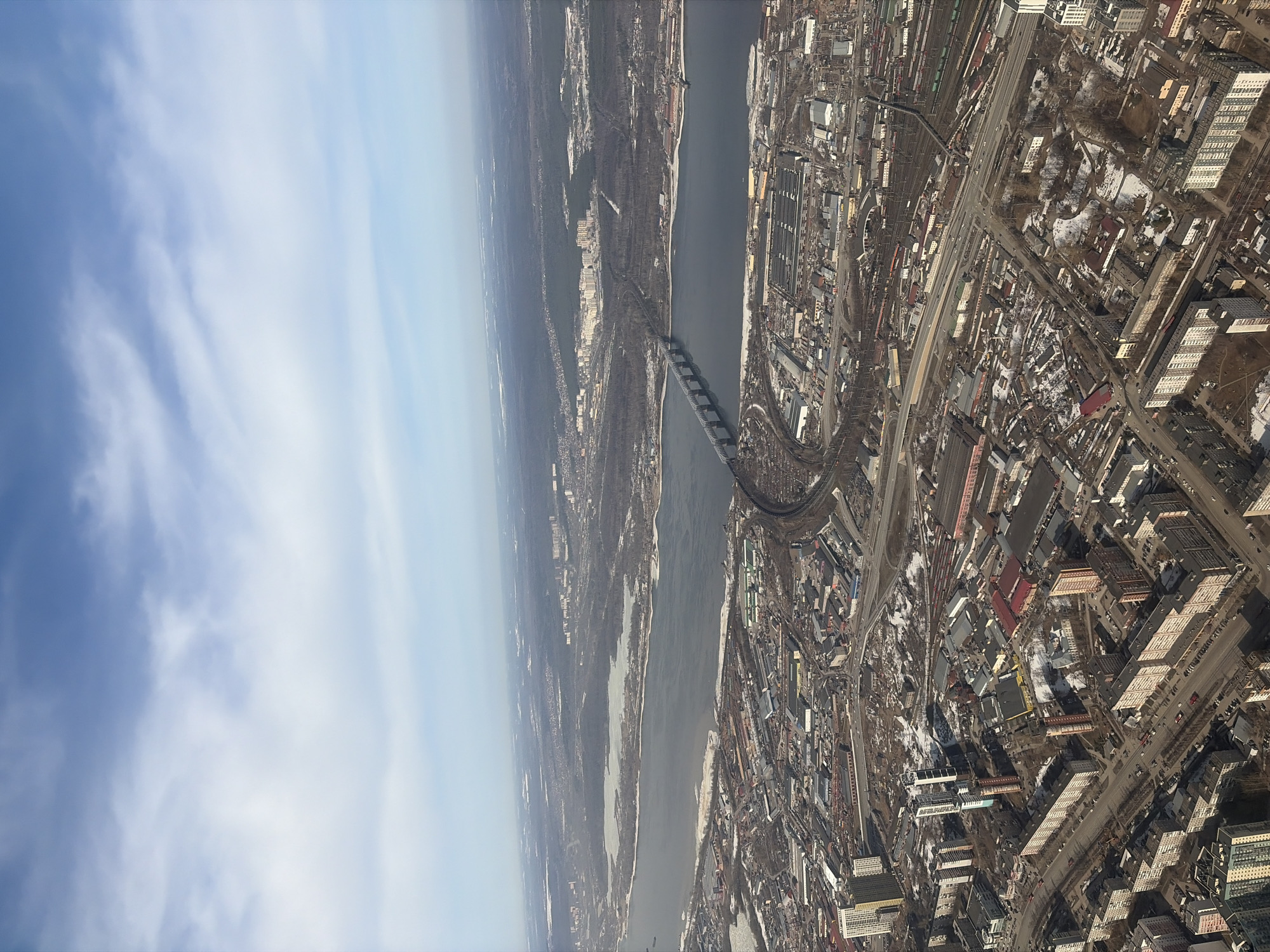 This aerial view shows the city of Perm, Russia, split by the Kama River. A bridge spans the river, connecting both sides of the city. The left side of the river appears less urbanized, with patches of forest and snow, possibly marking the outskirts or a less developed area. In contrast, the right side is densely built, with numerous buildings, roads, and infrastructure. There are some areas with snow patches among the buildings. The sky is partly cloudy, suggesting a fair weather day.