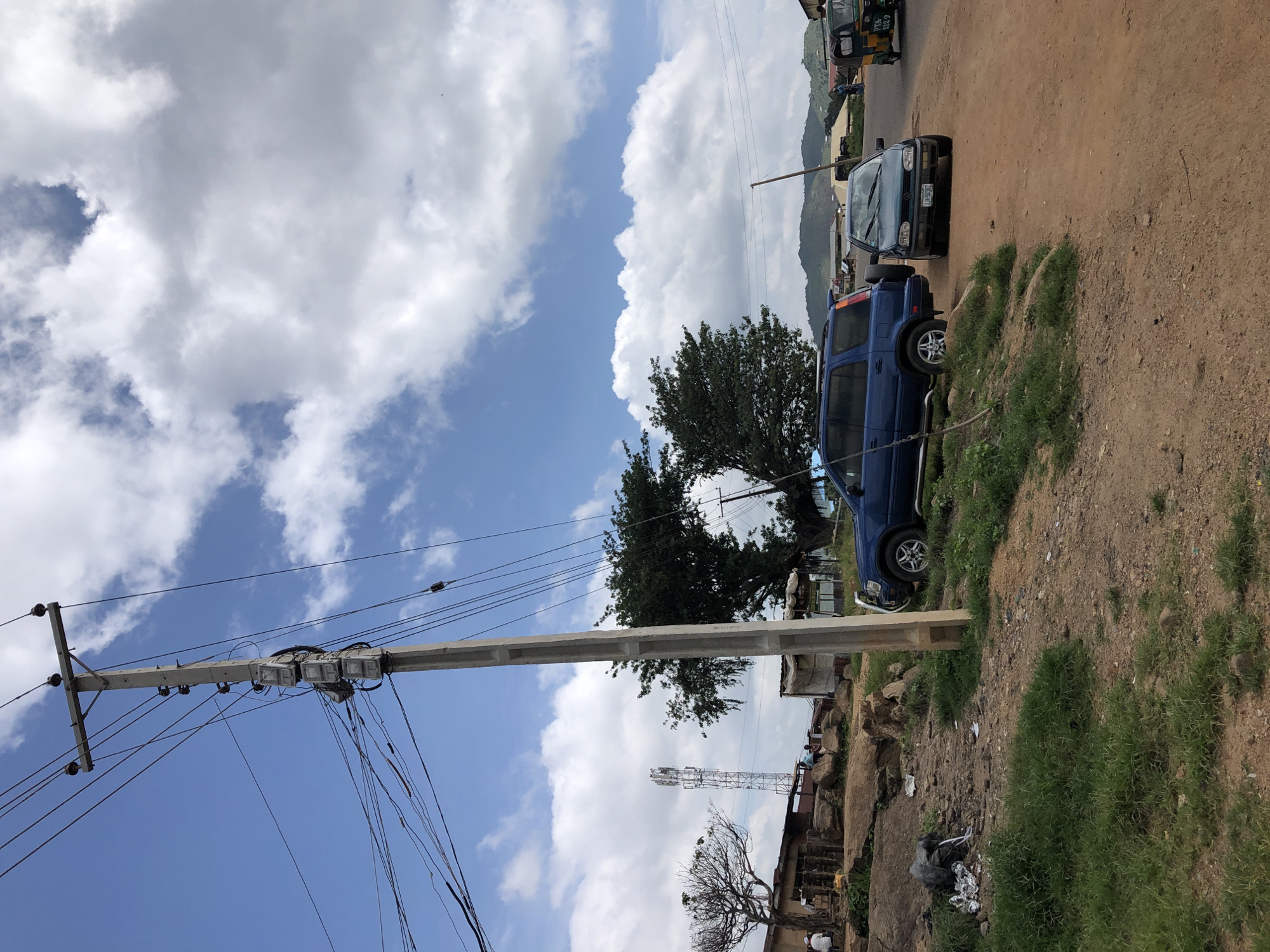 The image depicts a street scene in Jos, Nigeria, characterized by a concrete utility pole laden with numerous electrical wires, dominating the left side of the frame against a backdrop of a blue sky with scattered white clouds. A blue SUV is parked on the dirt road to the right of the utility pole, partially obscuring a smaller, dark-colored car. In the middle ground, a cluster of buildings and rocky terrain are visible, with a tall communications tower in the distance. A sparse, bare-branched tree stands near the buildings. The foreground is a dirt path with patches of green grass. The overall impression is of a somewhat dusty, unpaved urban environment.