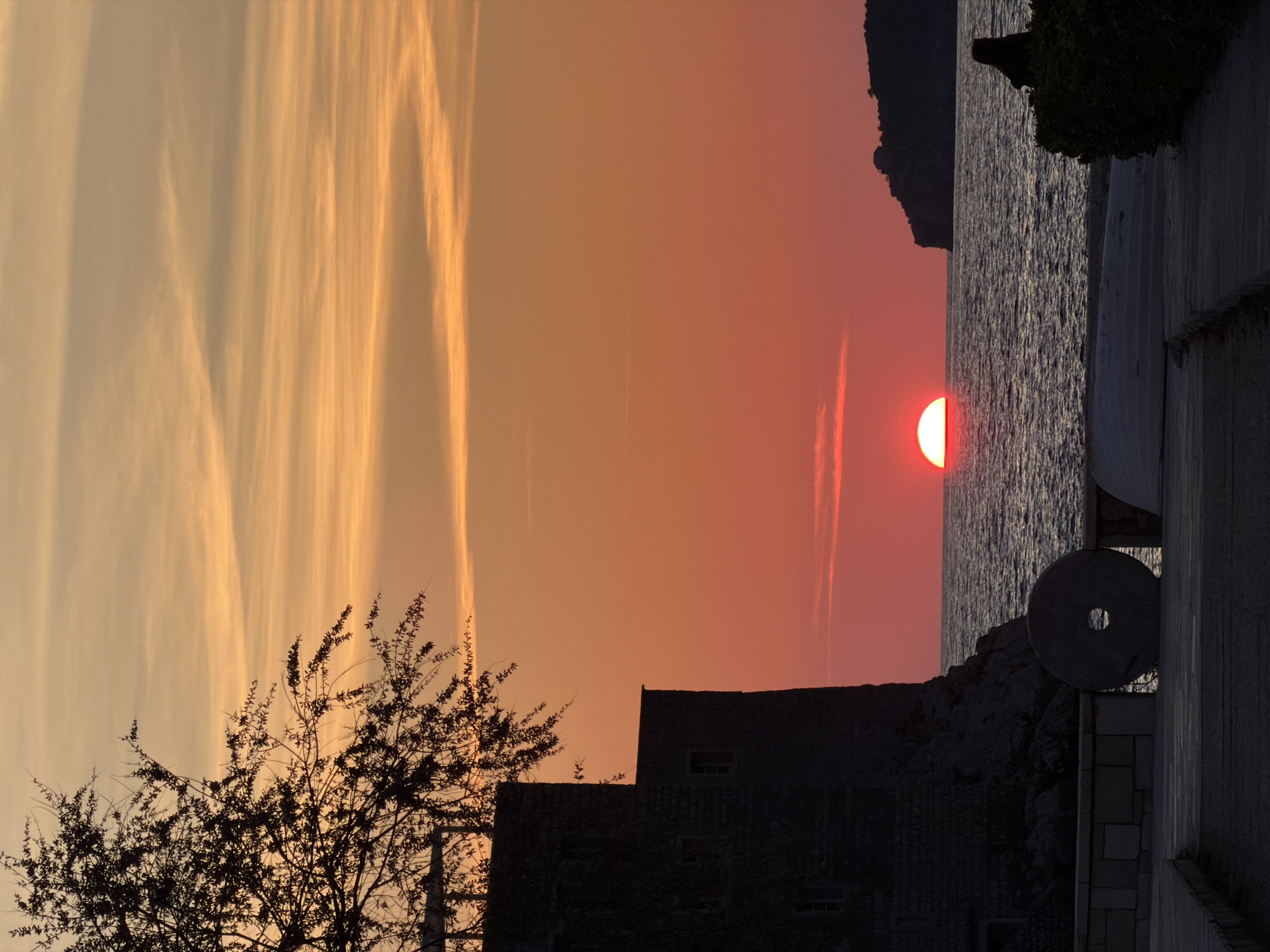 The image depicts a coastal sunset scene in Sveti Stefan, Montenegro. The sun, a prominent red and white half-disk, is partially submerged on the horizon line, centered above a dark blue-grey sea with visible ripples. The sky transitions from a pale orange-yellow at the top to vibrant orange and deep red towards the horizon, marked by horizontal streaky clouds.
On the far left, a dark, silhouetted stone building with small windows rises from the mid-ground, its surface appearing textured. A bare-branched tree or shrub, heavily silhouetted, partially covers the upper left sky and building. In the lower-left mid-ground, a large, dark circular stone object with a central hole, resembling a millstone, rests upright on a low stone wall beside the building.
To the right of the sun, a dark, rugged island or peninsula is silhouetted against the horizon, with an indistinct structure visible on its peak. In the lower right, dark bushes partially obscure two elongated, light-colored objects on a paved surface, which could be small boats or covers. A dark, partially silhouetted human figure, likely seated or crouching, is visible behind these bushes, facing the sea. The foreground consists of a dark, paved stone walkway or terrace with stepped sections, leading towards the water.