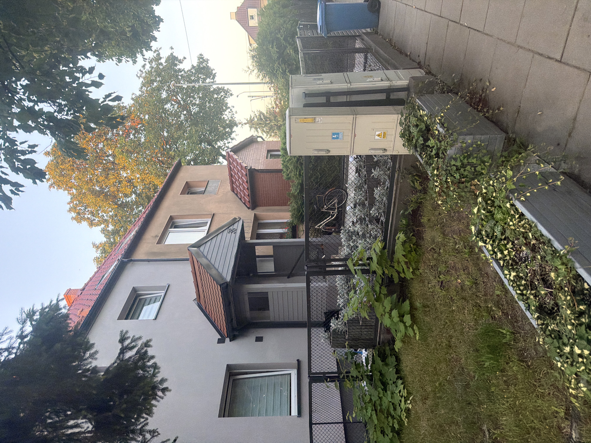 The image captures a residential street scene in Szczecin, Poland, with two adjoining buildings as the primary focus. The building on the left is a two-story structure with a light gray facade and a red tile roof. It features a prominent entrance with a grey porch roof and a door. A dark chain-link fence runs along the front of the property, separating it from the sidewalk. In front of the fence, there is a patch of green grass and several planters filled with lush green foliage.
The building to the right is a slightly taller structure with a tan-colored facade and a red tile roof. It shares a similar architectural style with the neighboring building. In the yard of this building, a white bicycle is visible leaning against the fence. Further to the right, on the sidewalk, are two stacked electrical utility boxes with warning labels. Next to these boxes, a blue trash bin is positioned near the fence. The sidewalk itself is paved with gray square tiles. Overhead, the branches of mature trees, some with golden-tinged leaves, create a natural canopy, suggesting an autumn setting. The sky is a pale blue, indicative of early morning or late afternoon light. There are no people visible in the image, and no discernible actions are taking place. The scene is static and peaceful.
