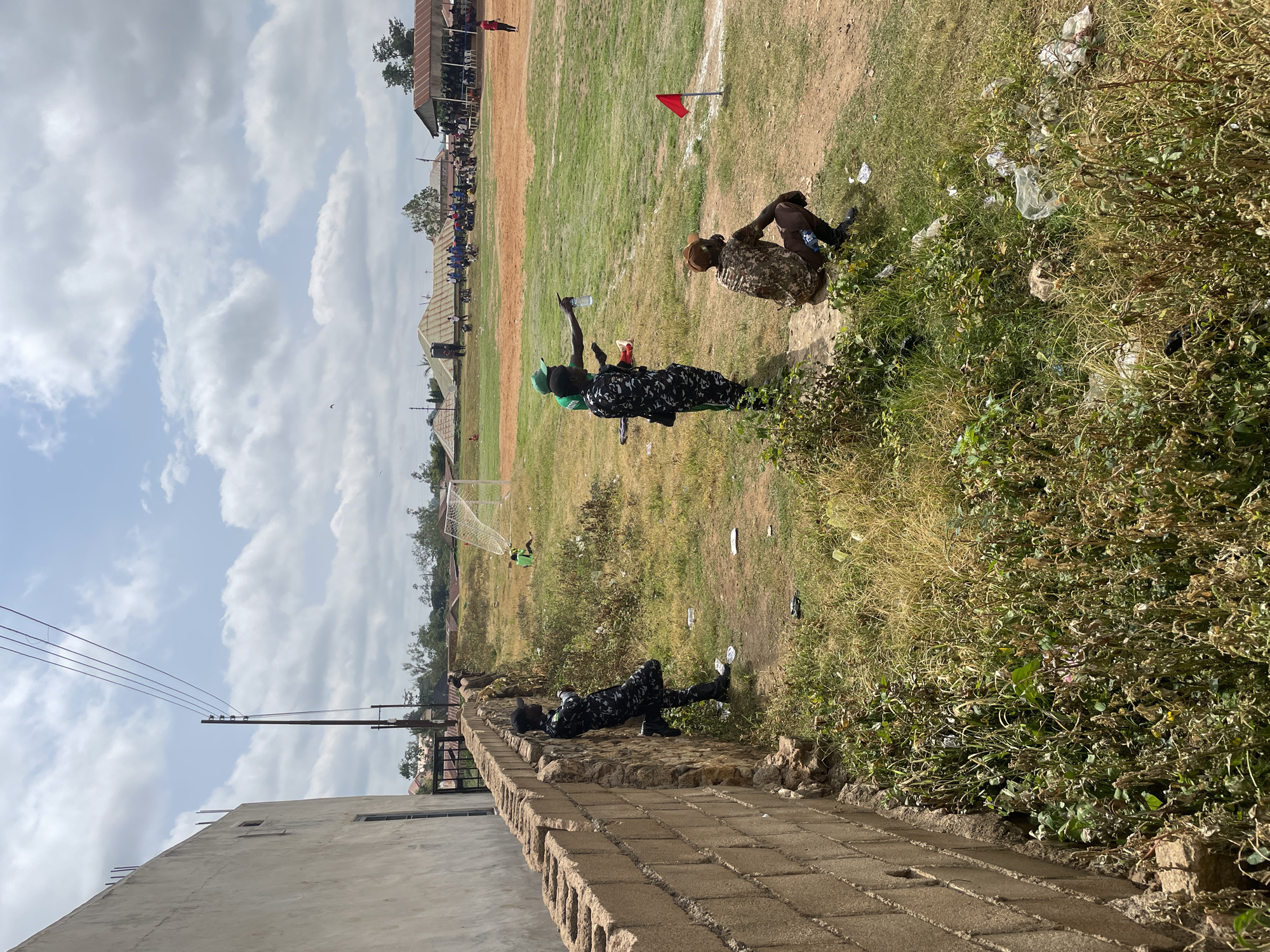 In Bukuru, Nigeria, this image captures a scene on a dusty, open field, likely a sports ground, with a clear sky and scattered clouds. In the foreground, a rough, brick wall runs along the left side, with overgrown dry grass and debris at its base. To the right of the wall, three individuals are present. One, wearing a black cap and camouflage uniform, leans against the wall, looking down. Further into the field, two other men in camouflage uniforms stand, one pointing towards the distance. A third man, wearing a camouflage shirt and brown trousers, squats nearby. In the background, a soccer goal and players can be seen on the field. A small gathering of people, some under a shaded structure, are visible further in the distance, along with several buildings and structures with corrugated iron roofs. The overall environment appears to be an outdoor event or gathering in a semi-rural or developing urban area.