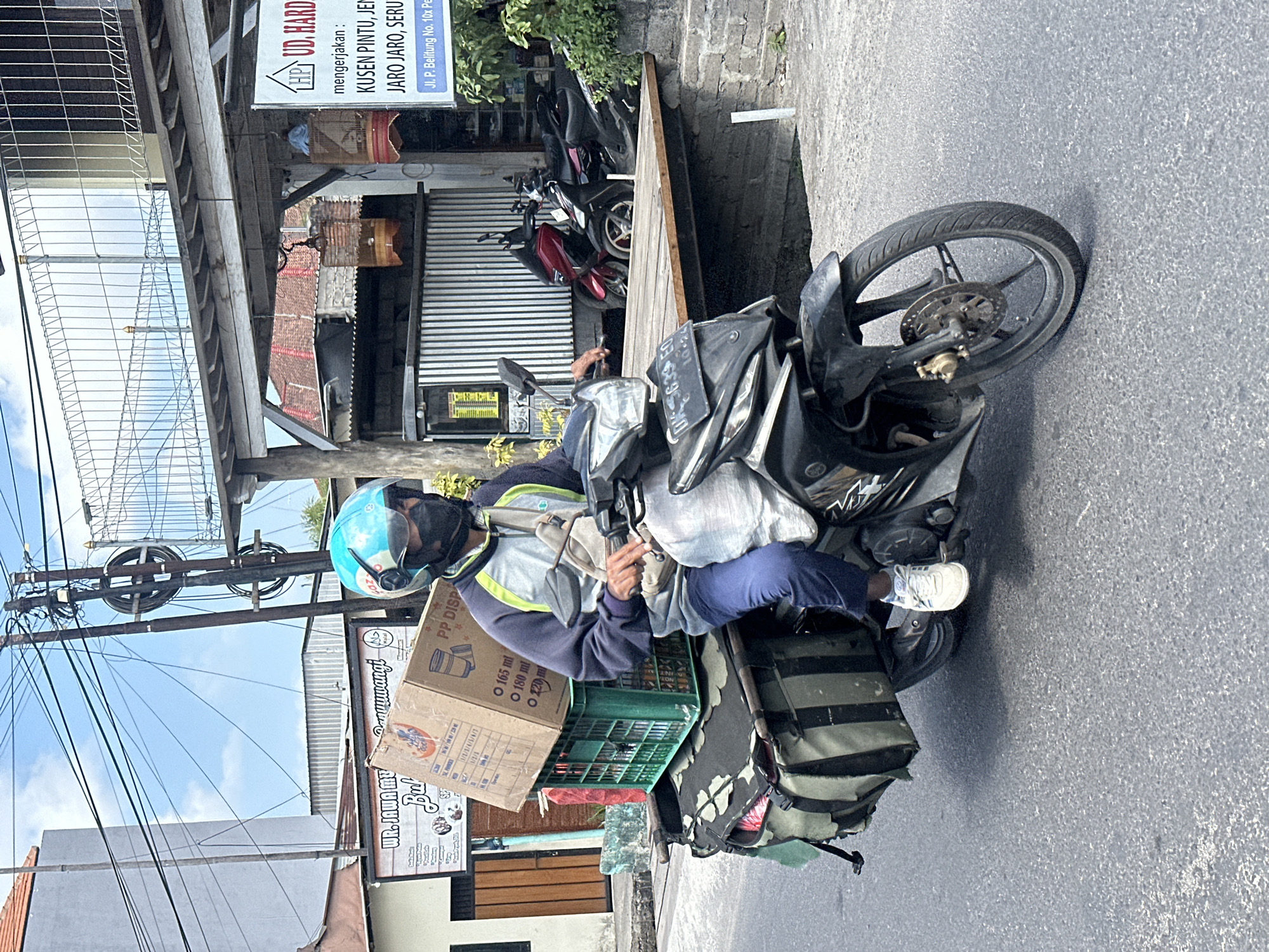 A person wearing a blue helmet and a black face mask rides a motorcycle on a street in Denpasar, Indonesia. The motorcycle is loaded with items, including boxes and green crates. The person is wearing a blue and gray jacket, blue pants, and white sneakers. In the background, there are buildings and a sign that reads "UD. HARD." The street is paved, and there are power lines overhead. Two other motorcycles are parked in the background. The weather appears to be clear with a blue sky.