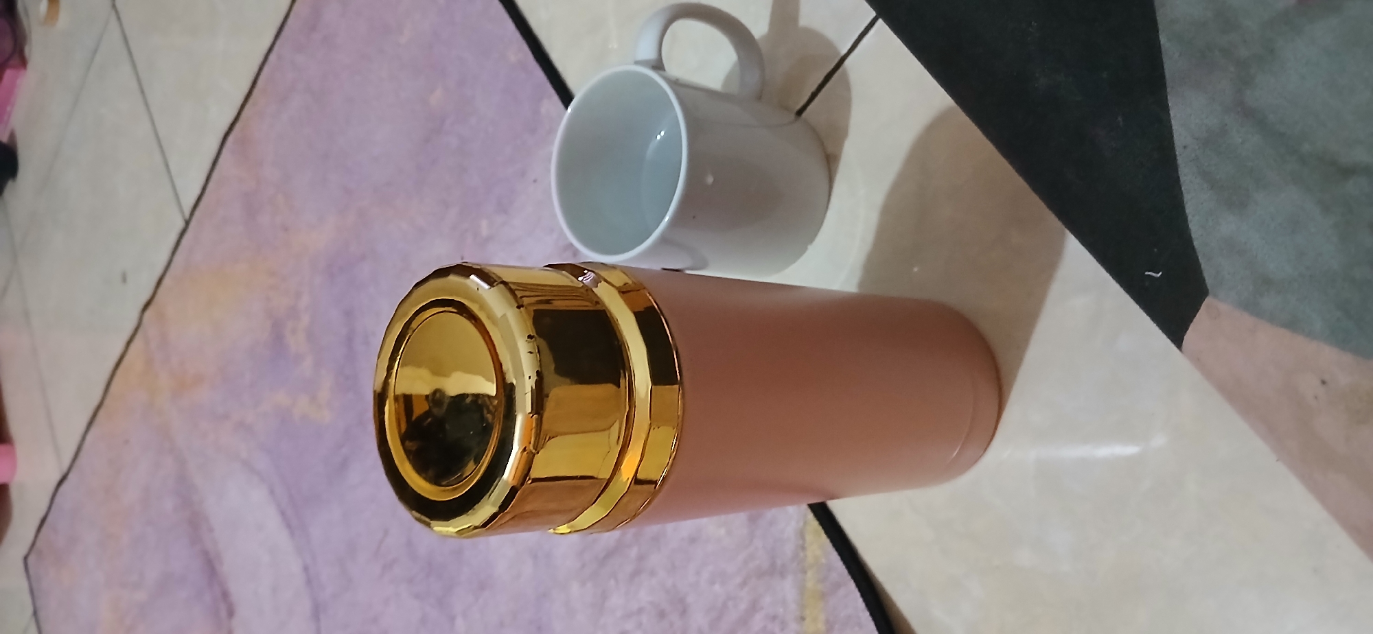 The image shows a pink and gold thermos and a white mug on a tiled floor with a purple rug in the background. The scene is indoors, likely in a home setting. There are no people visible in the image, and no specific landmarks are depicted that would indicate its location in Samarinda, Indonesia. The objects are stationary, and no actions or interactions are occurring. The lighting appears to be natural or artificial indoor lighting.