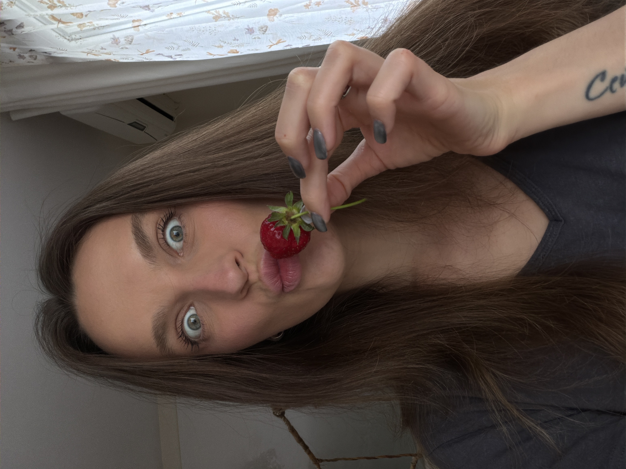 A person, likely a female, is holding a strawberry to her lips with her right hand. Her eyes are wide and she appears to be making a playful expression. The person has long brown hair, fair skin, and a tattoo on her left forearm. The fingernails are painted gray. The background shows a window with light-colored floral curtains and an air conditioning unit. The location cannot be confirmed to be Budva, Montenegro, using only the visual information from the image.