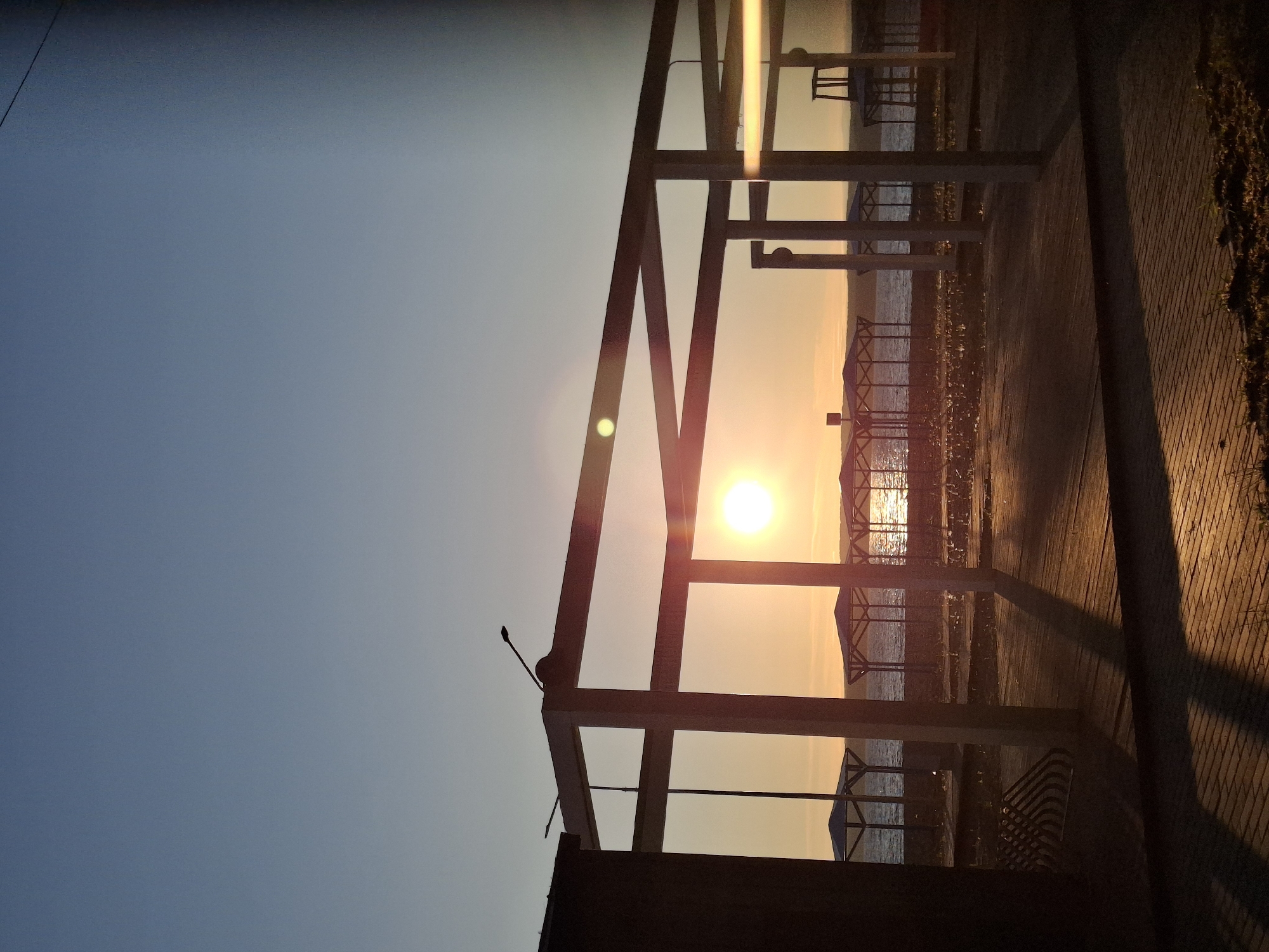 The image depicts a sunset over the Kama River in Chaikovsky, Russia. The scene is viewed from what appears to be a promenade or pier, with a row of covered cabanas visible along the riverbank. In the foreground, there is a paved walkway and a wooden deck. The sun is low in the sky, casting long shadows and reflecting on the water. The sky is a gradient of blue and orange. There are no people visible in the image. The overall impression is one of a quiet, serene evening by the river.